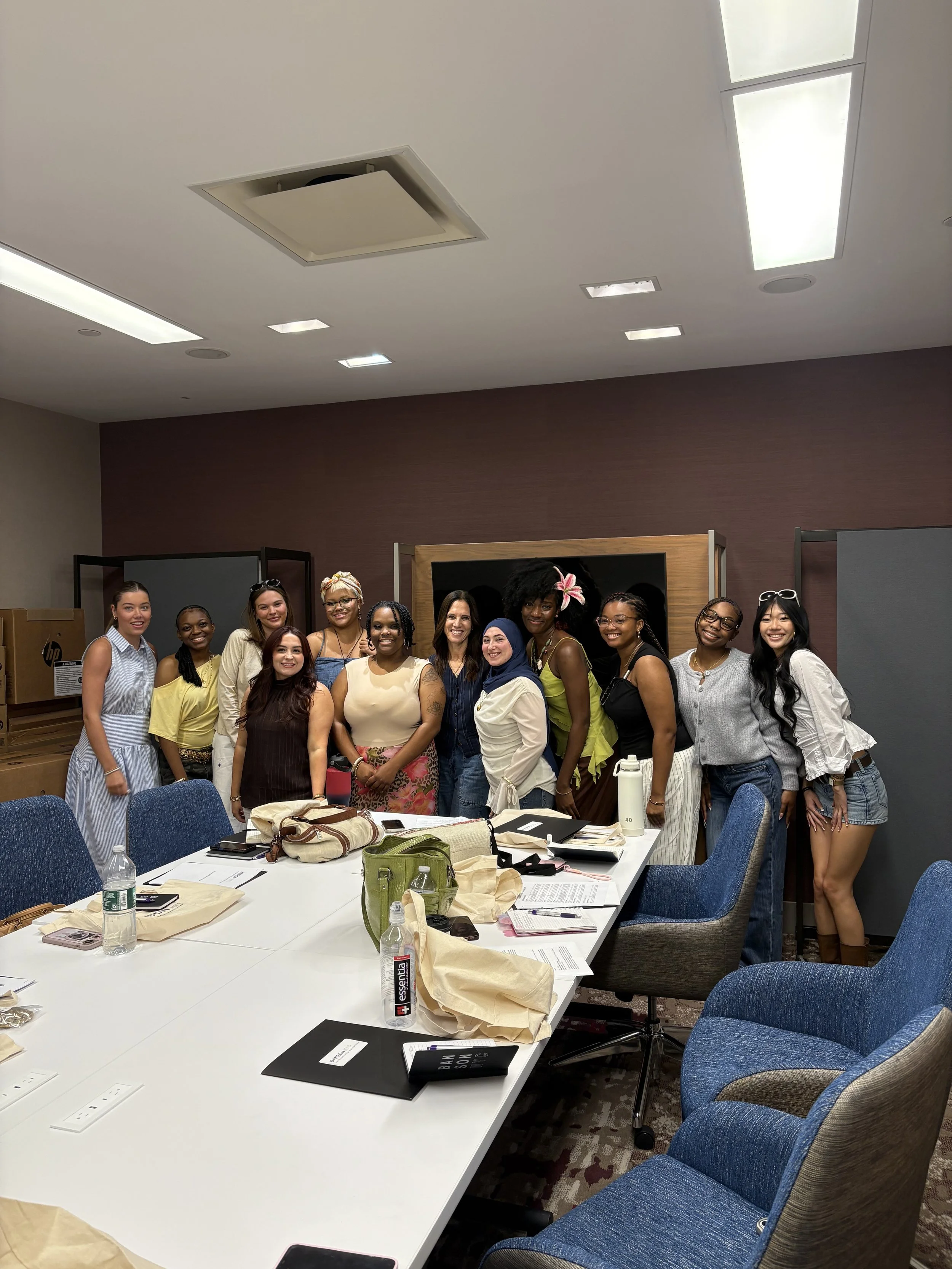A group of diverse women and a man posing for a photo inside a conference room with tables, chairs, and various personal items on the table.