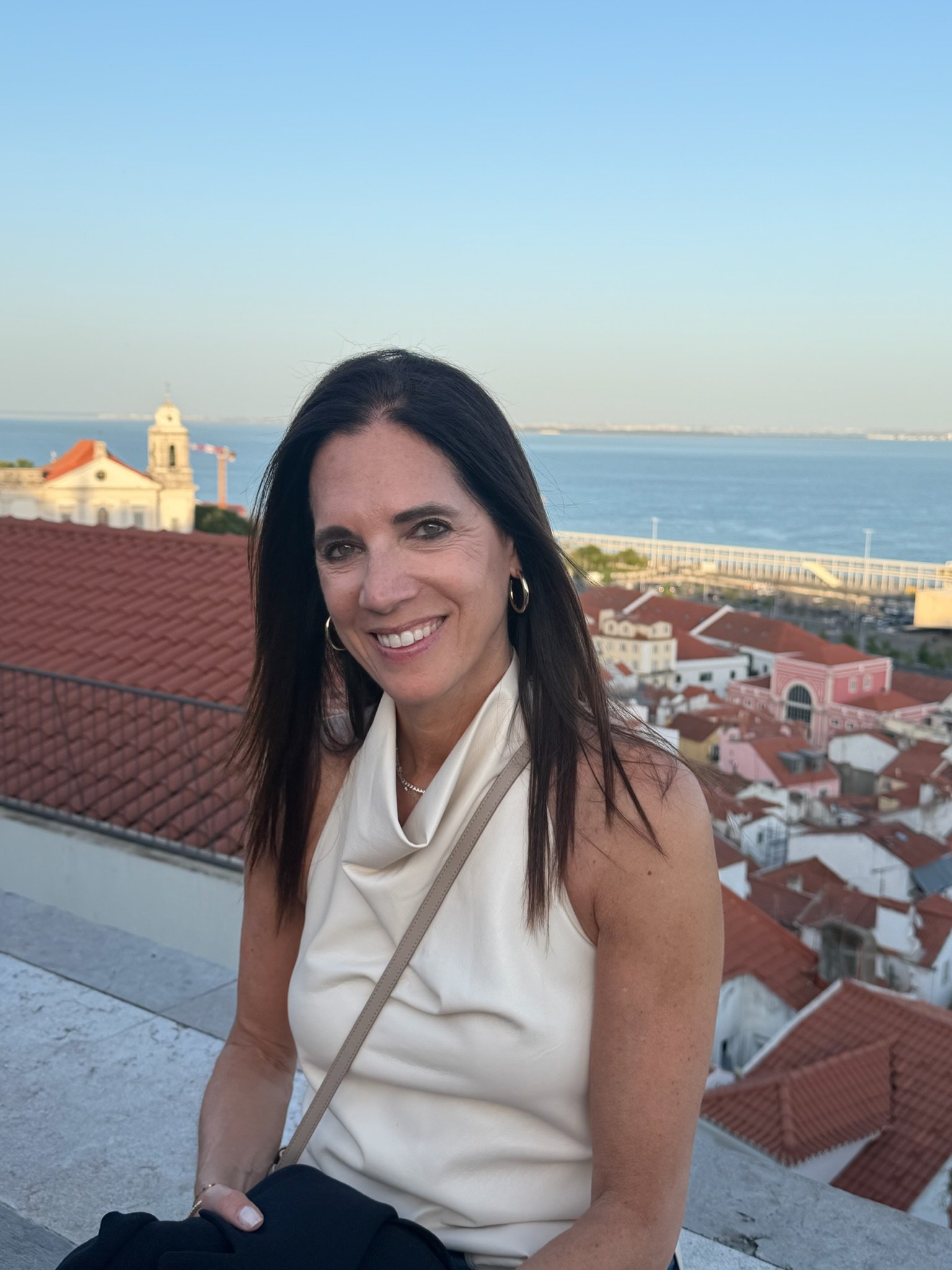 A woman smiling outdoors near a coastal area with rooftops, a church, and the ocean in the background.