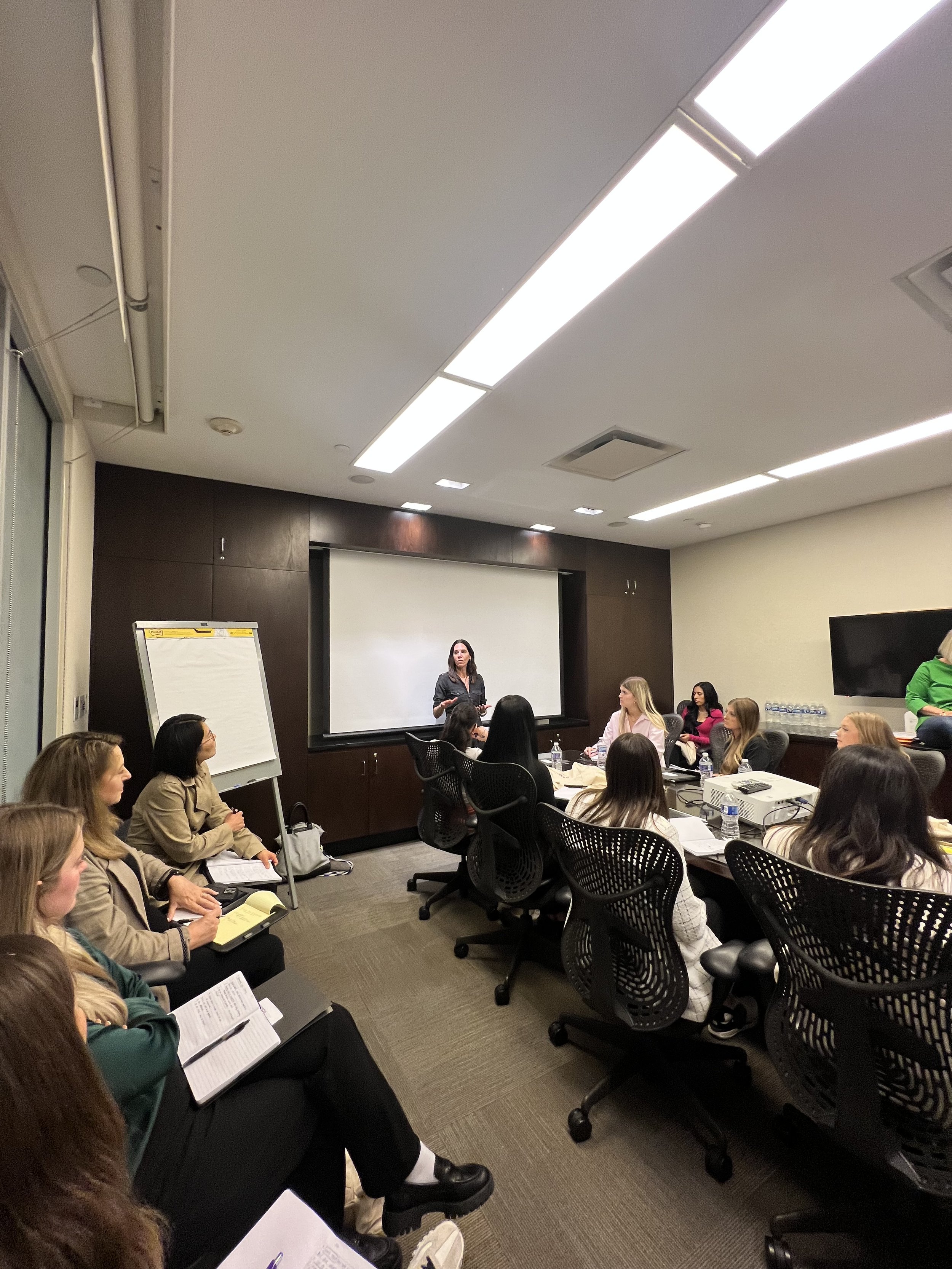 A woman is standing in front of a group of seated women in a conference room, giving a presentation. The room has a large white screen behind her, with ceiling lights and a whiteboard nearby.