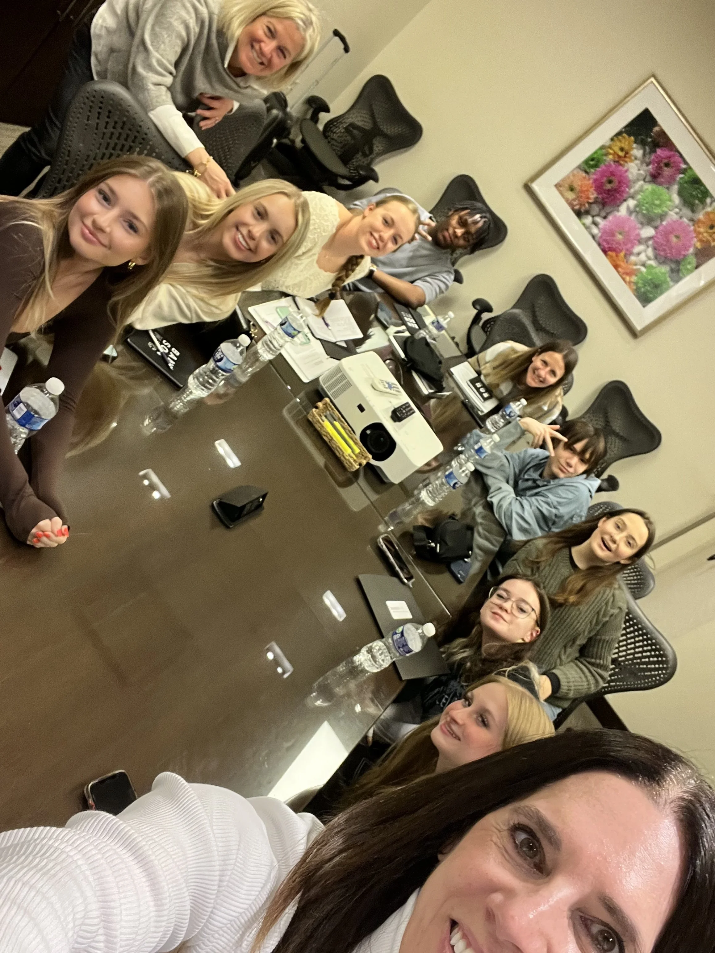 Group of women smiling and taking a selfie in a conference room decorated with a framed floral picture.