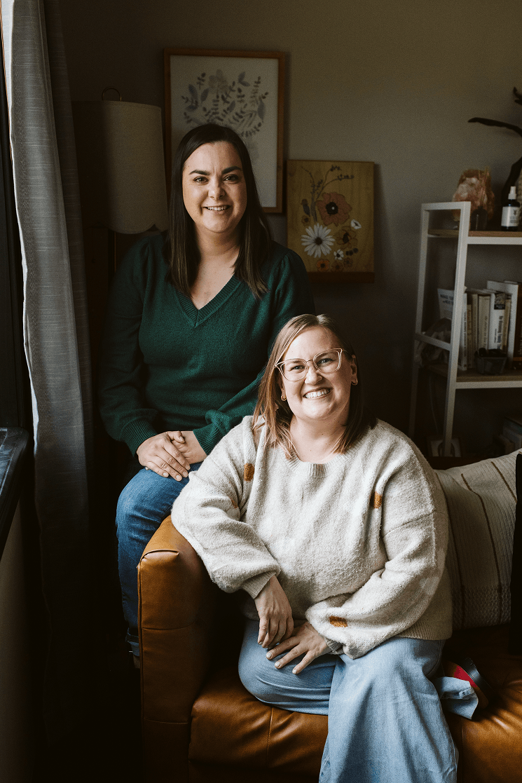 Two women smiling indoors, one sitting on a leather armchair and the other standing behind her, in a cozy room with artwork and books in the background.
