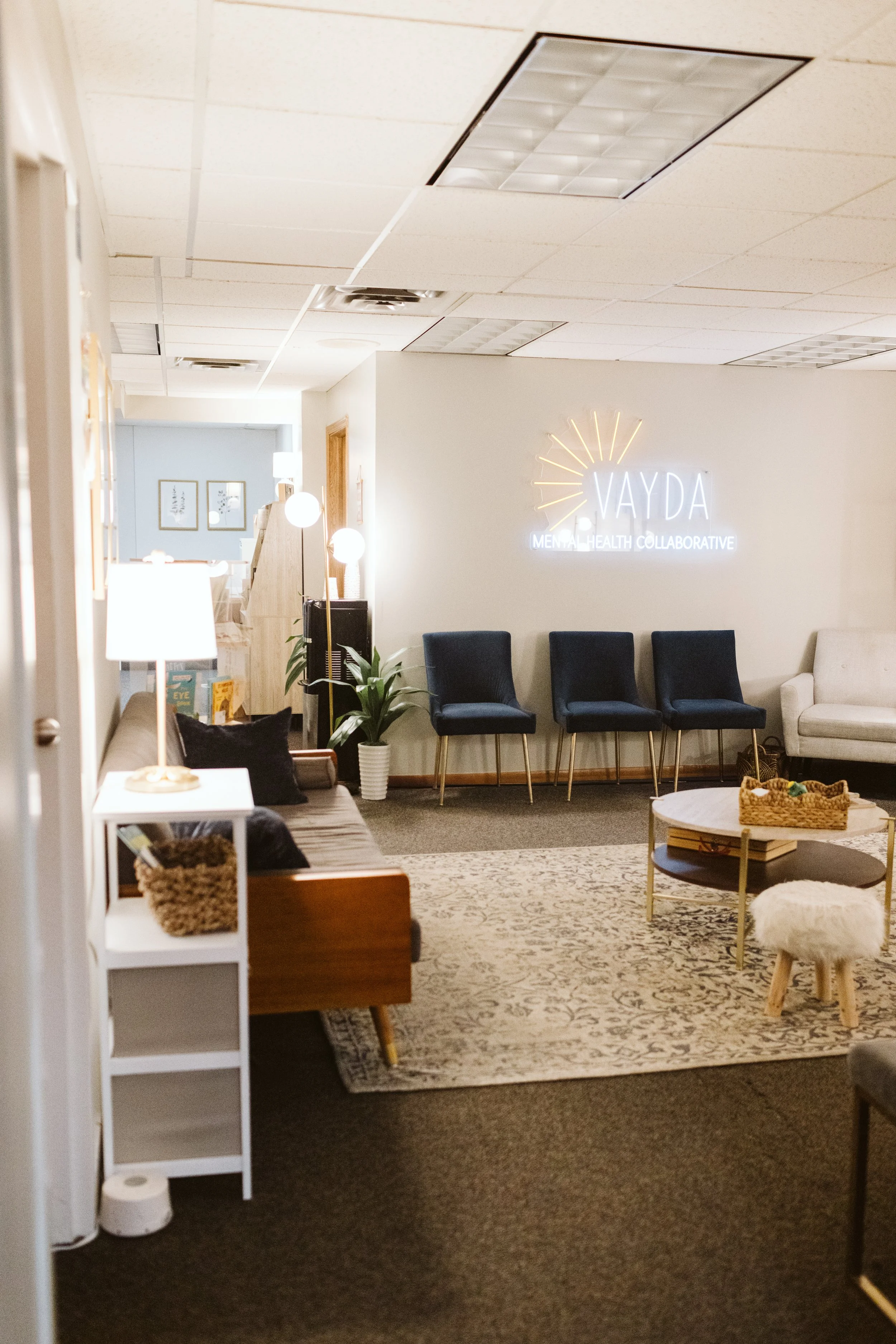 Interior waiting room with chairs, a side table, a rug, and a neon sign that reads 'VAYDA' with 'MENTAL HEALTH COLLABORATIVE' below. Decor includes framed artwork and table lamps.