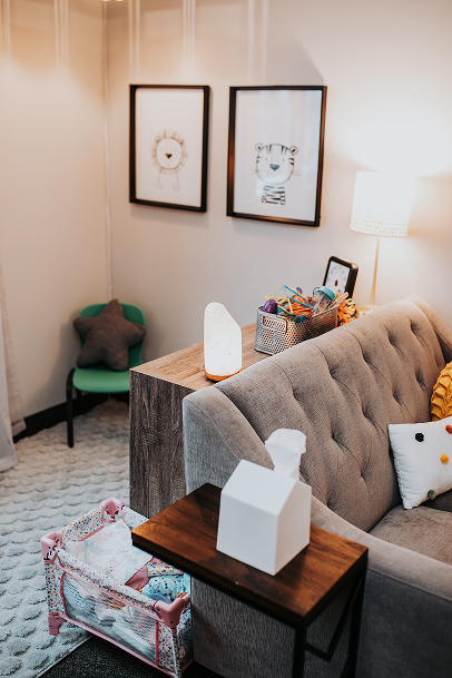 Living room with three framed animal art pieces on the wall, a grey tufted sofa with decorative pillows, a small side table with a tissue box, salt lamp, and a basket filled with toys, a green chair with a plush pillow, a white floor lamp, and a soft textured carpet.