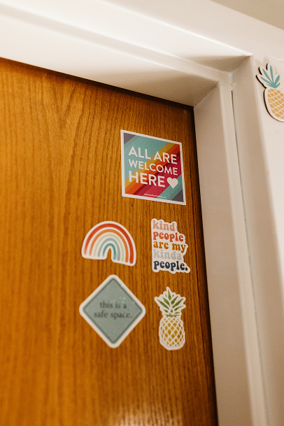 Decorative stickers on a wooden door, including a rainbow, a pineapple, and colorful text saying 'All are welcome here' and 'kind people are my kind people'.