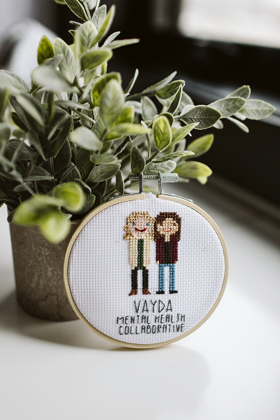 Cross-stitched embroidery hoop with two figures and text, placed next to a potted green plant on a white surface near a window.