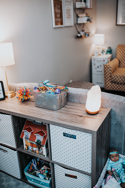 A living room with a wooden storage unit containing books and toys, a salt lamp, a small blue doll, and a colorful spiky ball. In the background, there is a patterned armchair, a white nightstand with a lamp, framed artwork and shelves with books on the wall.