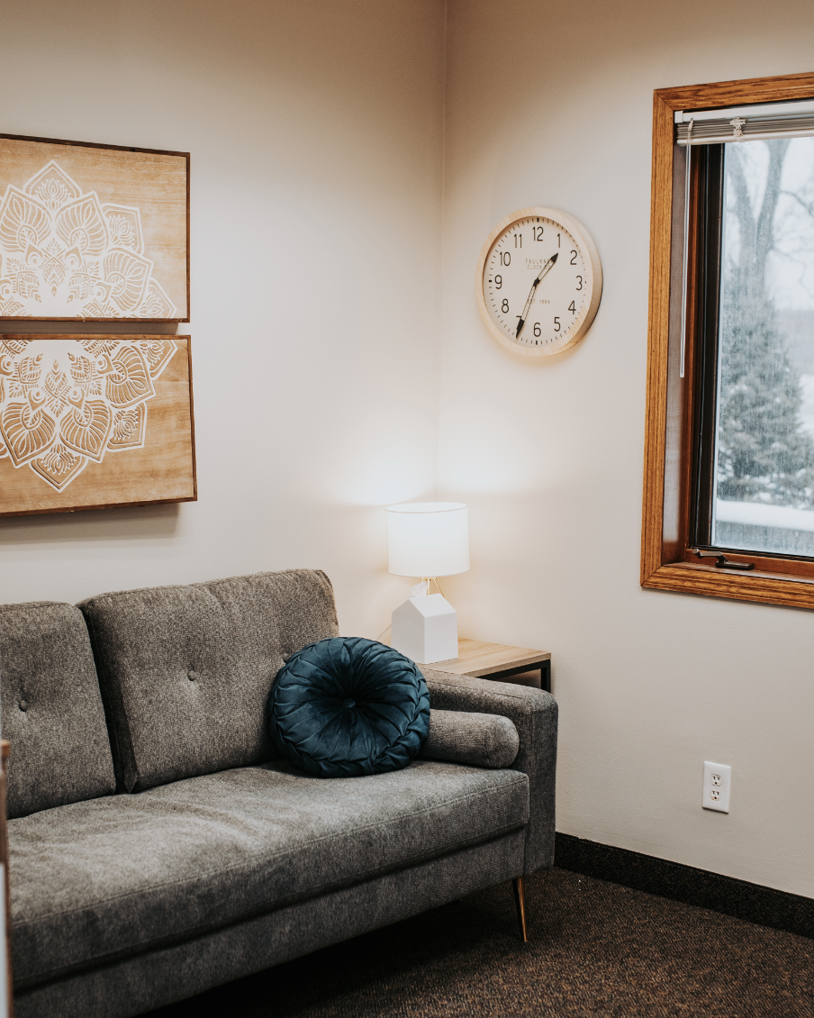 A cozy living room corner with a gray couch, a dark teal round throw pillow, a wooden side table with a white lamp, a wall clock showing 1:28, a window with a wooden frame, a white wall with a decorative wooden art piece, and an electrical outlet.