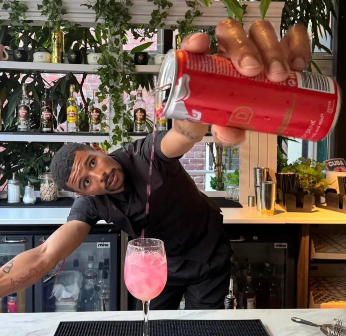 Barista pouring a pink drink into a glass with a bartender shirt, behind a bar with bottles and plants in a cafe or bar setting.