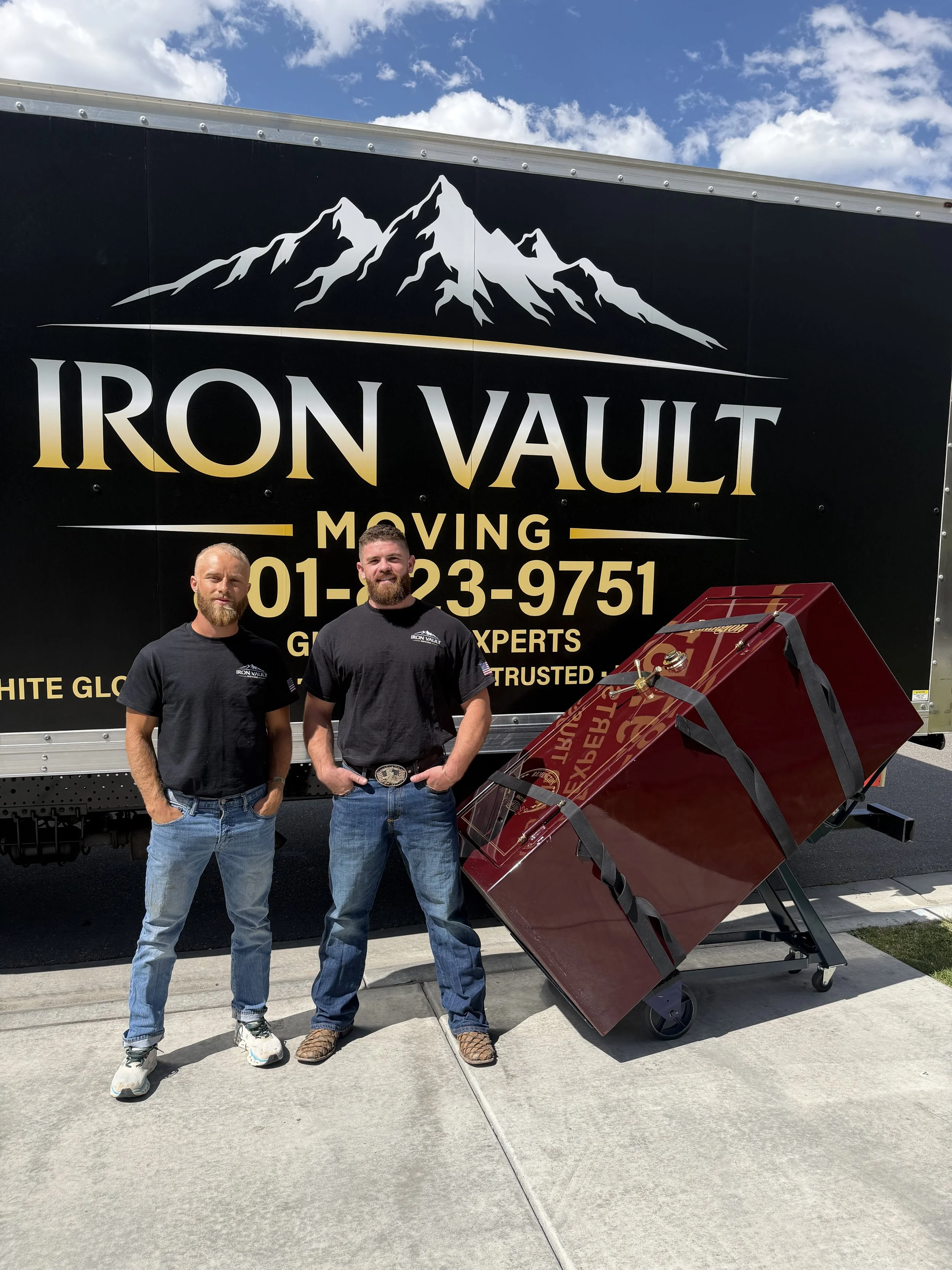 Two professional movers standing in front of custom box truck equipped to load and haul heavy items. "Iron Vault Moving" on the side of truck. next to them is a massive gun safe on a professional stair climbing dolly.