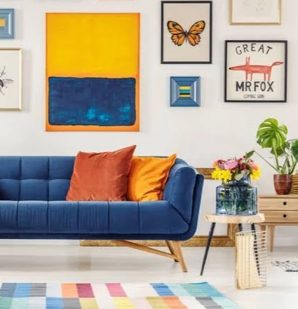 Salon avec un canapé bleu, coussins orange et jaune, mur décoré avec plusieurs tableaux, plantes vertes, et un tapis coloré.