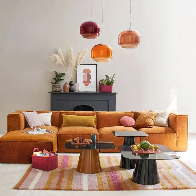 Salon de style moderne avec un canapé orange, plusieurs coussins, une table basse noire, un tapis coloré, et des suspensions en verre orange, rouge et rose