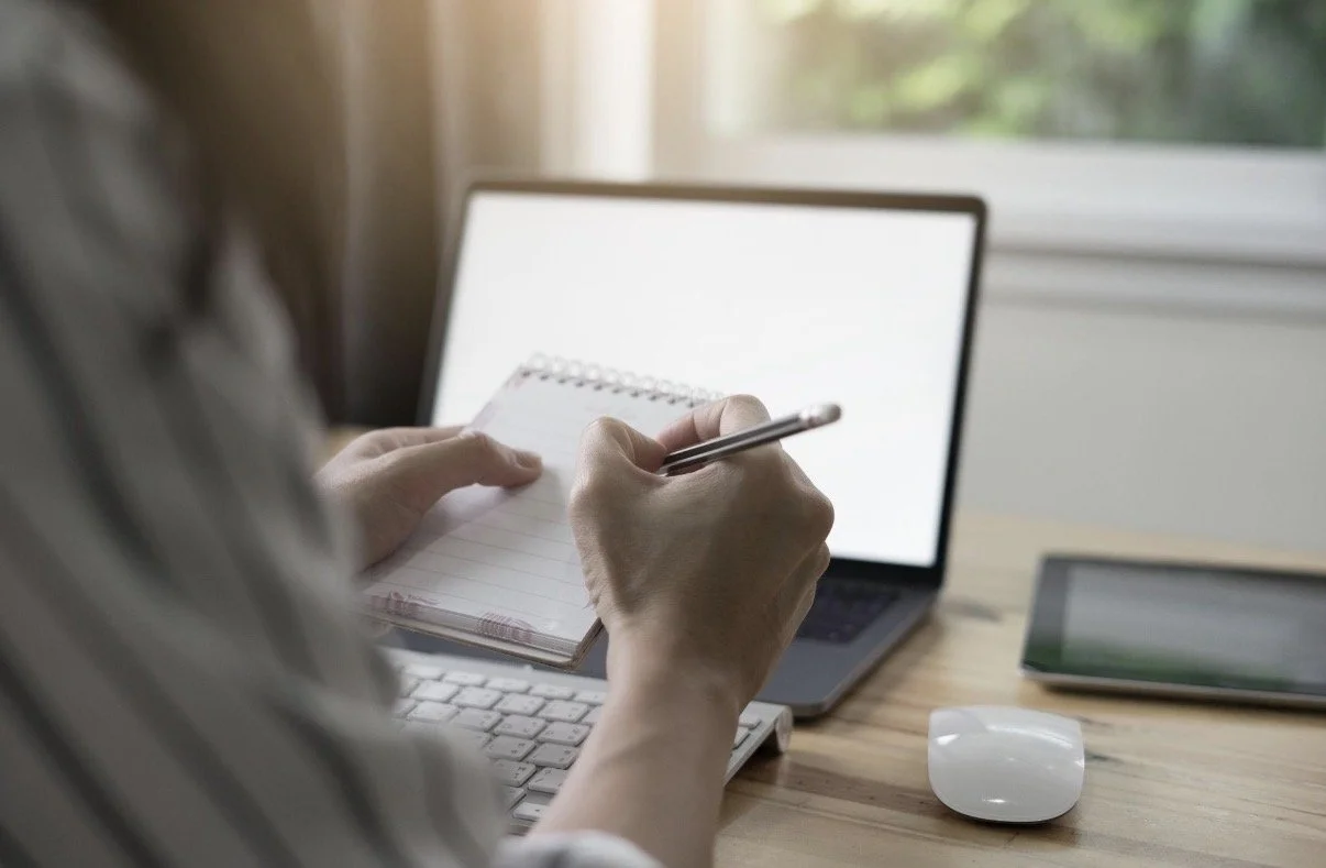 Person taking notes on a small notepad while working on their laptop at a desk, with a computer monitor, tablet, and mouse nearby.