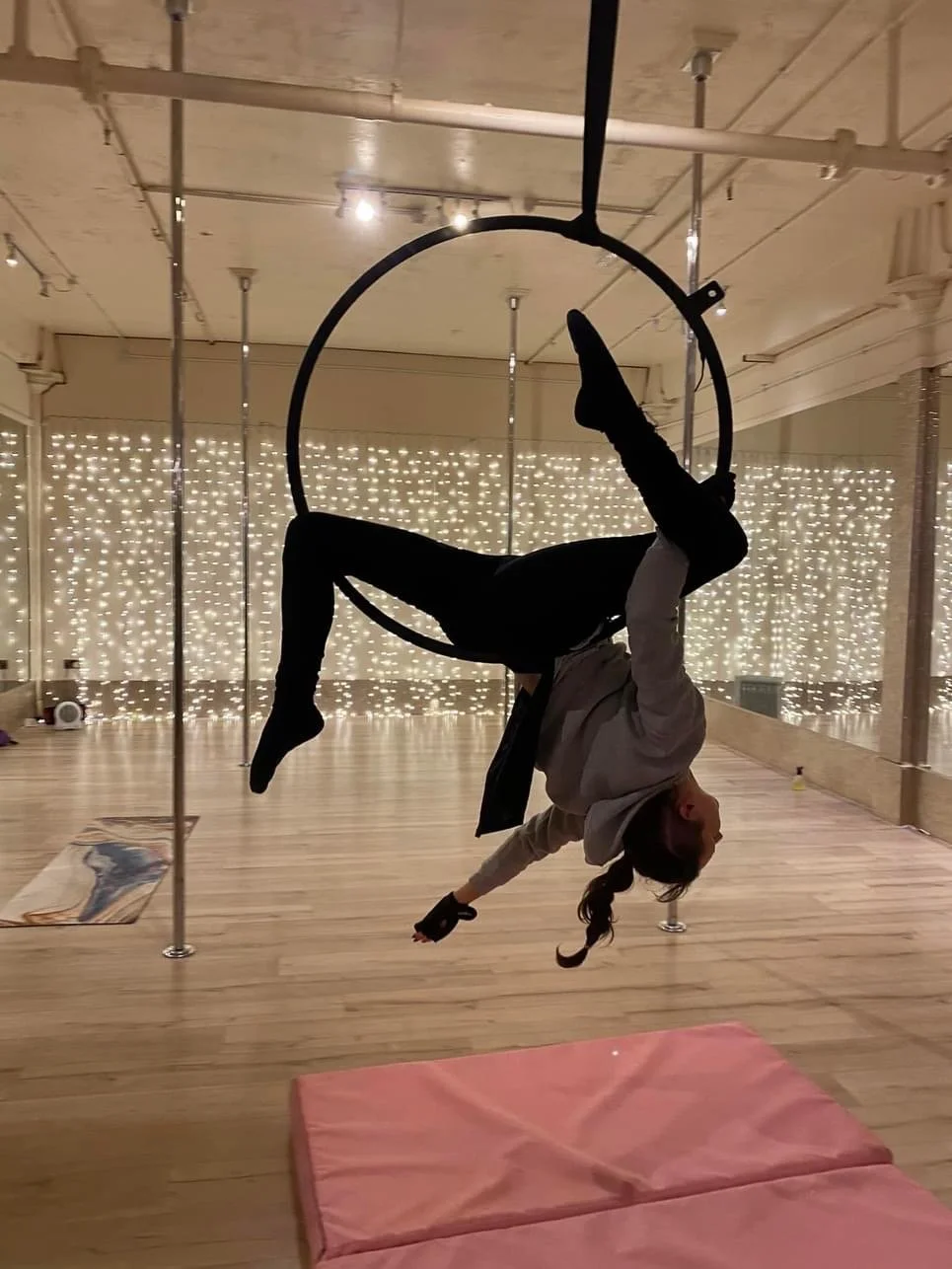 A person performing an aerial acrobatics routine on a hoop in a dance or fitness studio with wooden floors, soft lighting, a pink mat on the floor, and string lights on the back wall.