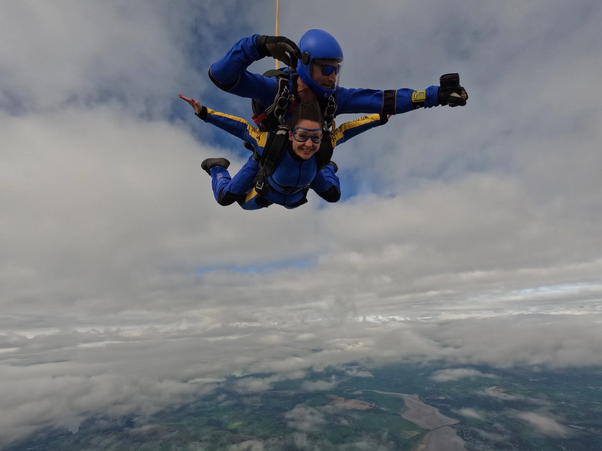 Two skydivers mid-air with clouds and blue sky in the background. The person in front is smiling and wearing goggles, while the person behind is holding onto them and wearing a helmet and sunglasses.