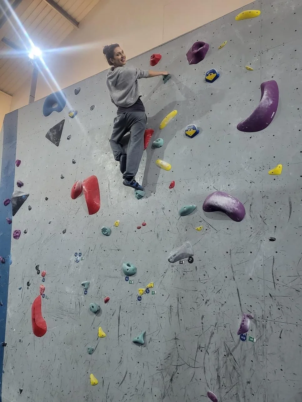 A young woman climbing an indoor bouldering wall with multicolored holds, wearing a gray sweatshirt and dark pants, smiling at the camera.