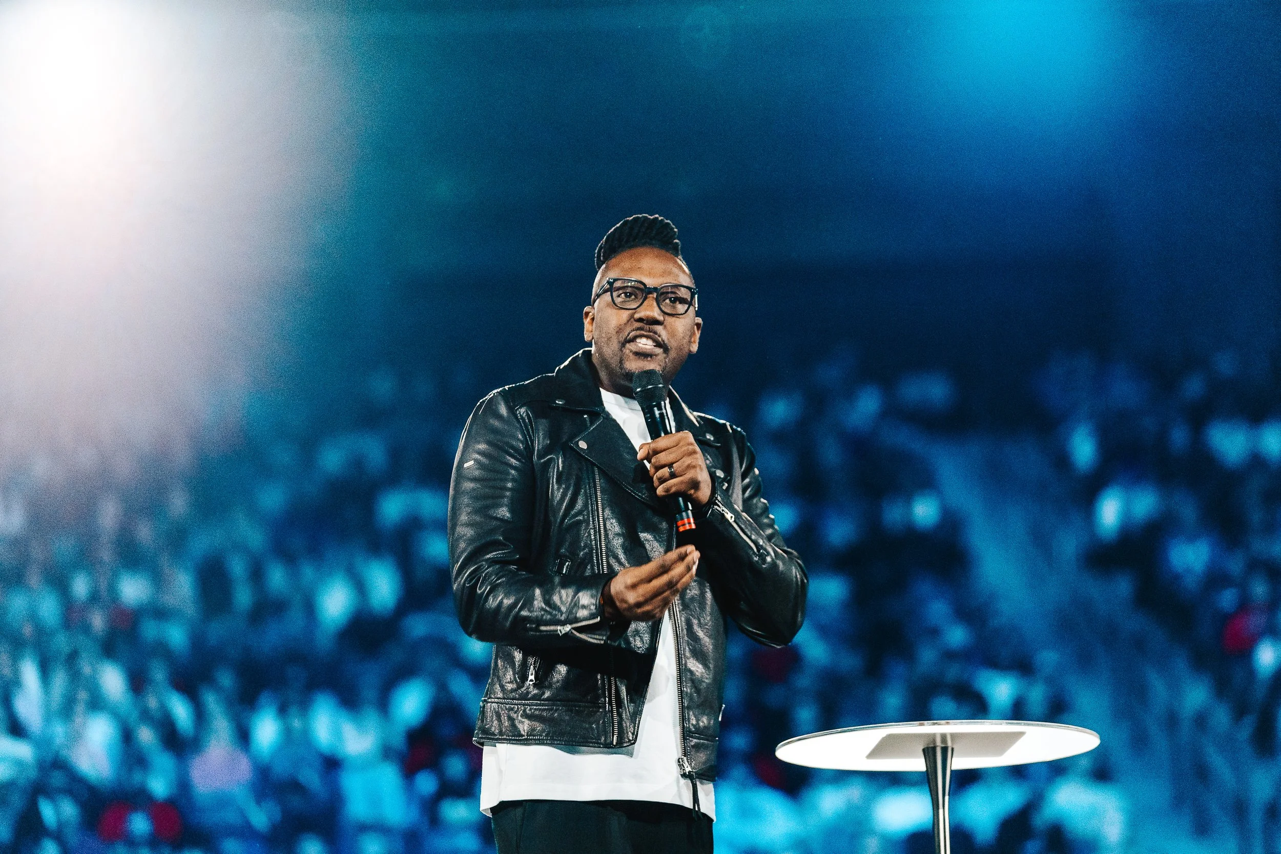A man with glasses and a beard, wearing a black leather jacket, holding a microphone and speaking on stage in front of a large audience, with a blurred crowd in the background.