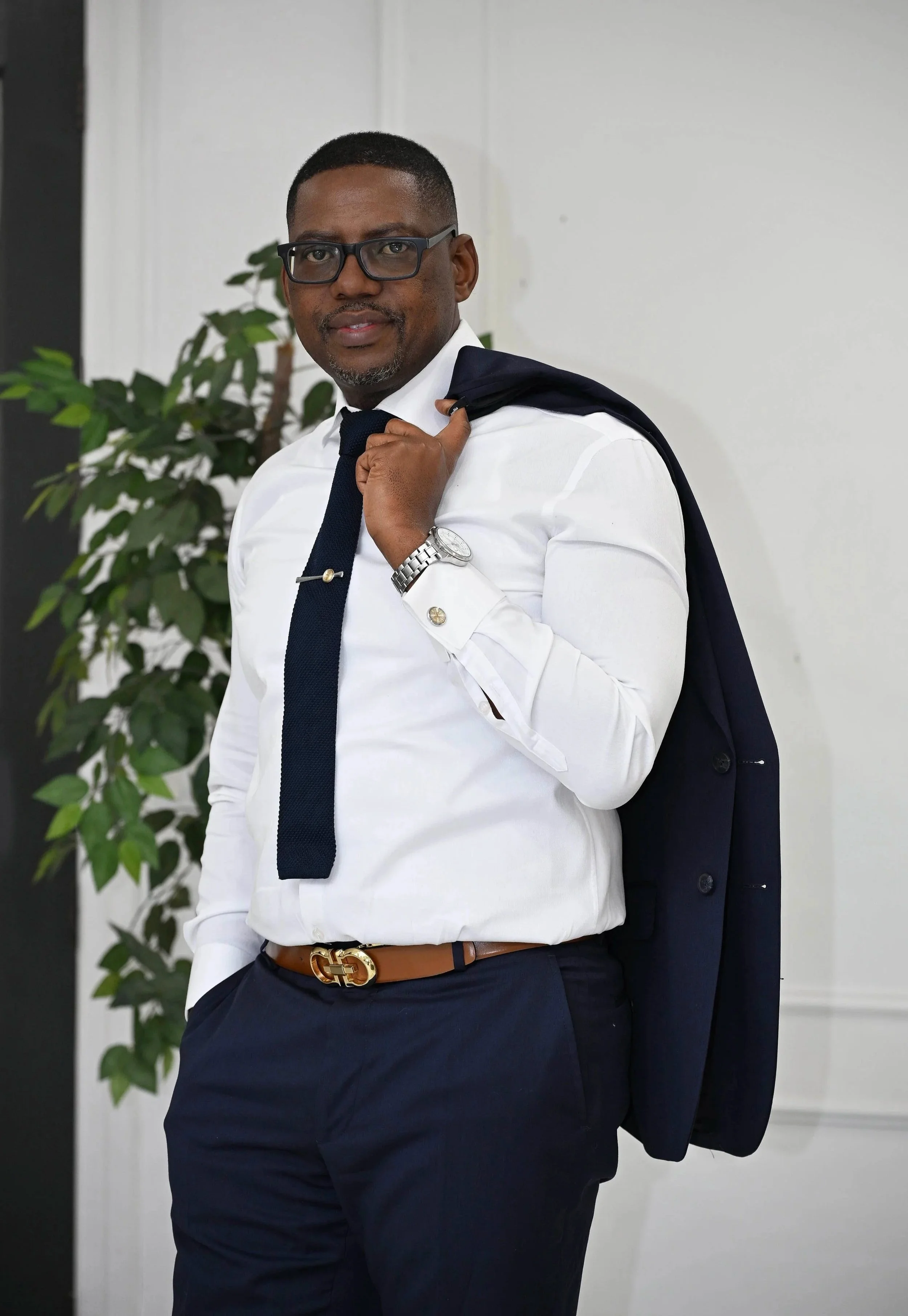 Man wearing glasses, white dress shirt, dark tie, tan belt, and navy pants, holding a navy blazer over his shoulder, standing inside next to a potted plant with green leaves.
