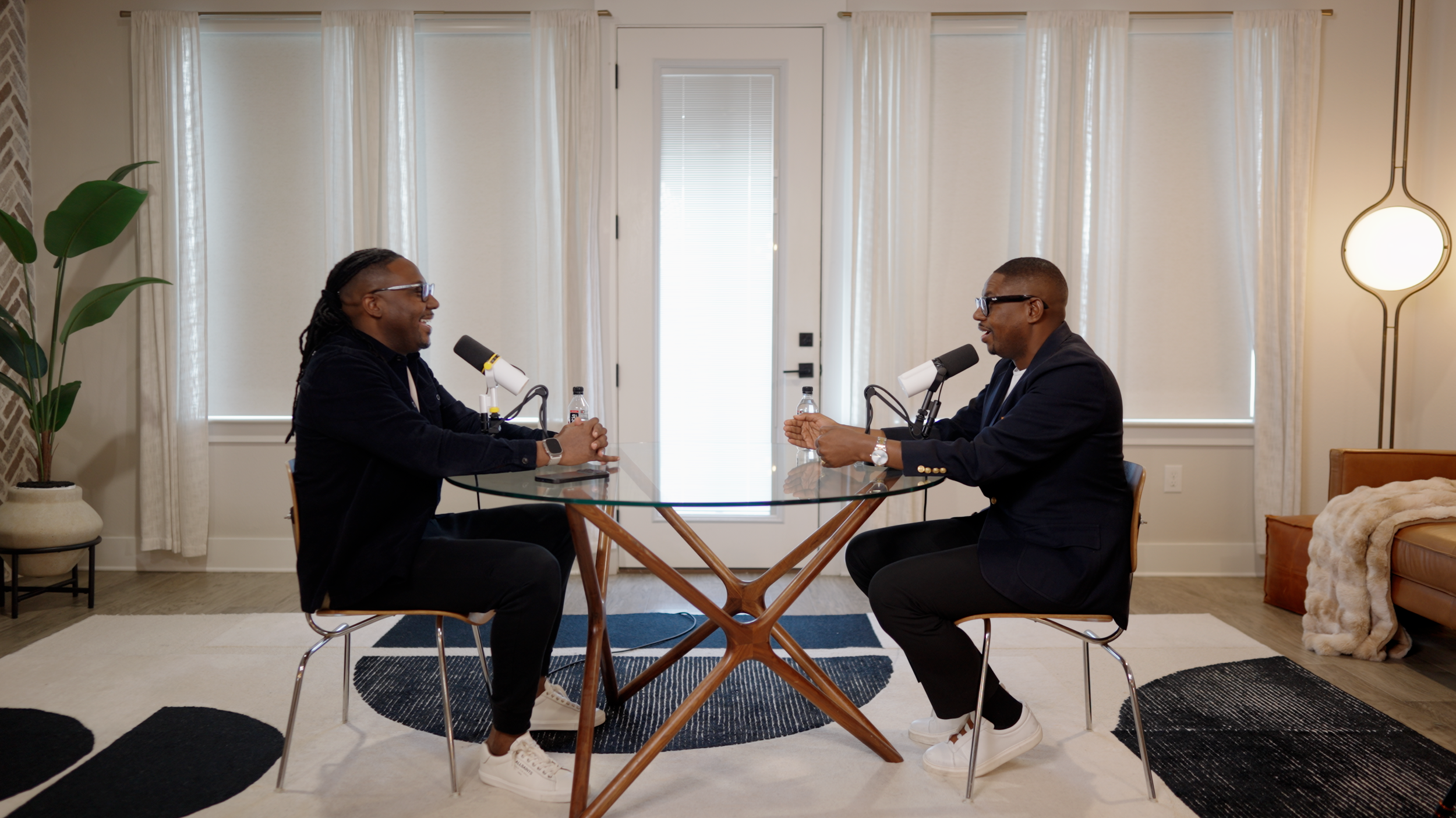 Two men sit across from each other at a round glass table, engaging in conversation, with microphones in front of them, in a bright room with large windows and off-white curtains.