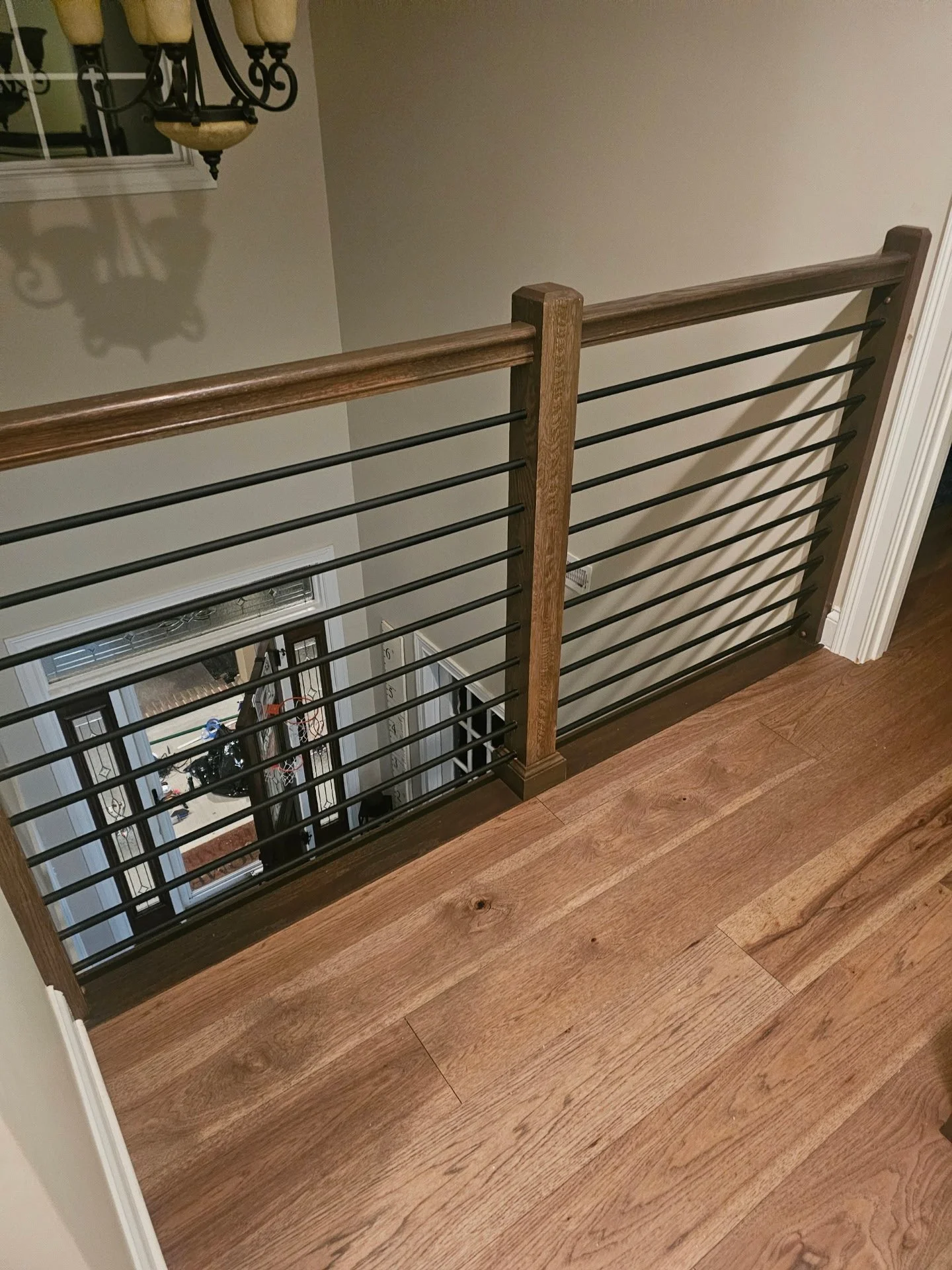 View of a staircase railing with wooden top and black metal bars, overlooking a lower level with a door and various items visible, next to a beige wall and hardwood floor.