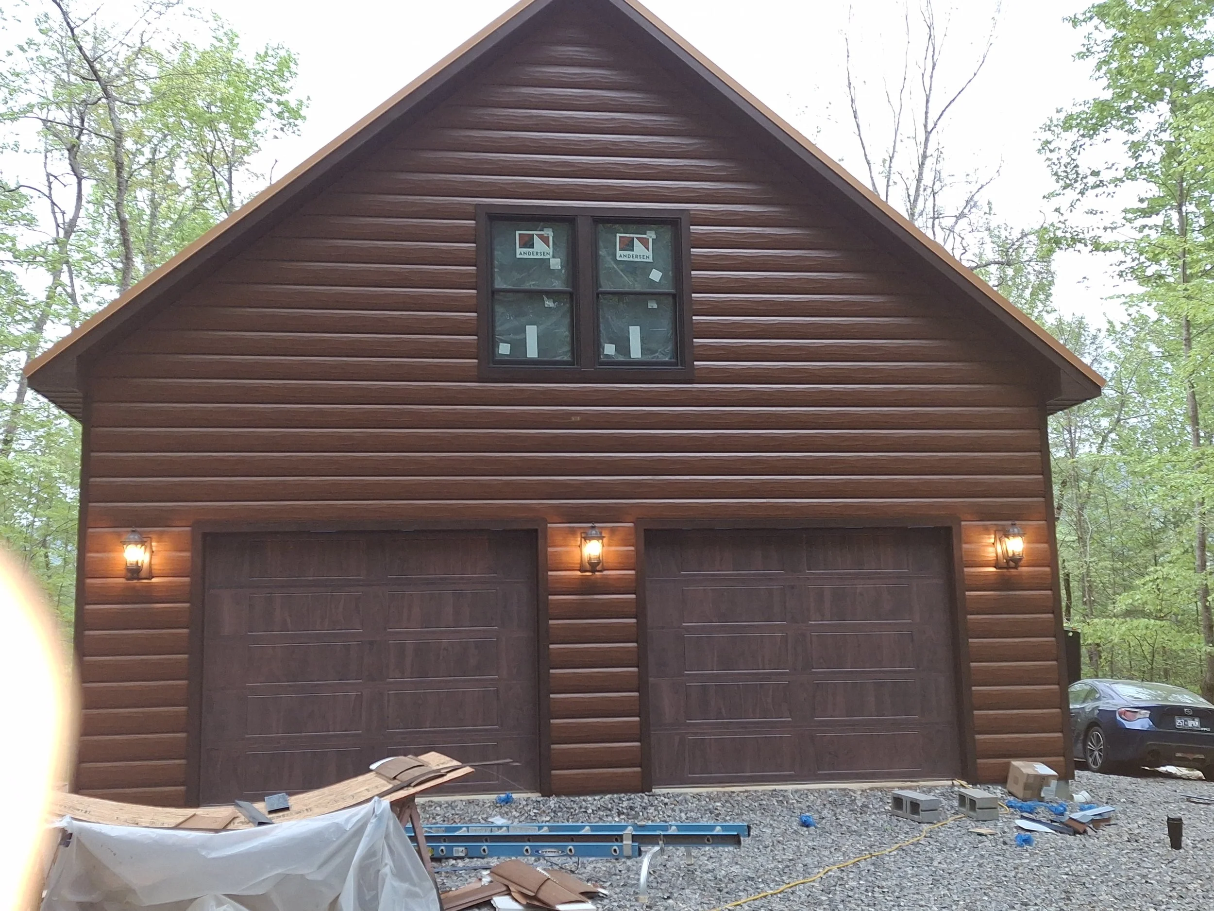 A two-story wooden house with a gable roof, brown siding, small window on the upper level with Andersen branding, and two garage doors on the lower level, illuminated by exterior lights, surrounded by trees, with construction materials and supplies i
