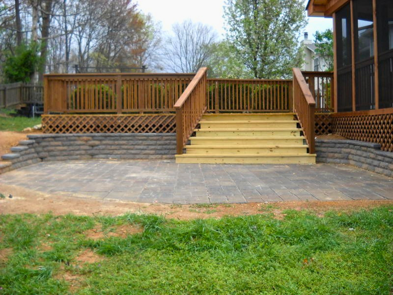 New wooden stairs leading up to a backyard deck with a railing, brick retaining wall, and a paved patio area. Grass in the foreground and trees in the background.