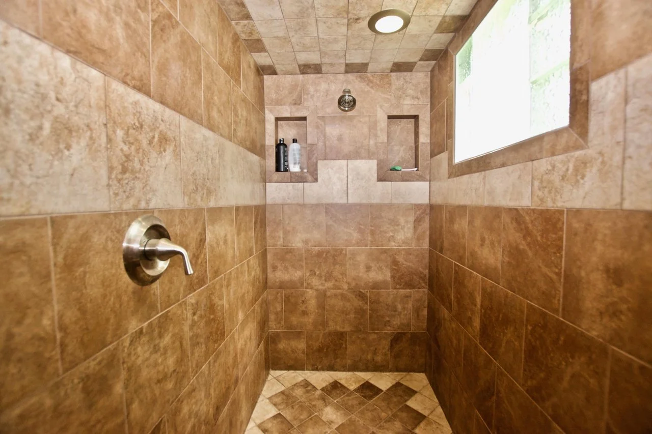 Interior of a tiled shower with brown and beige tiles, including built-in shelves with shampoo bottles, a window for natural light, and a metal shower faucet.