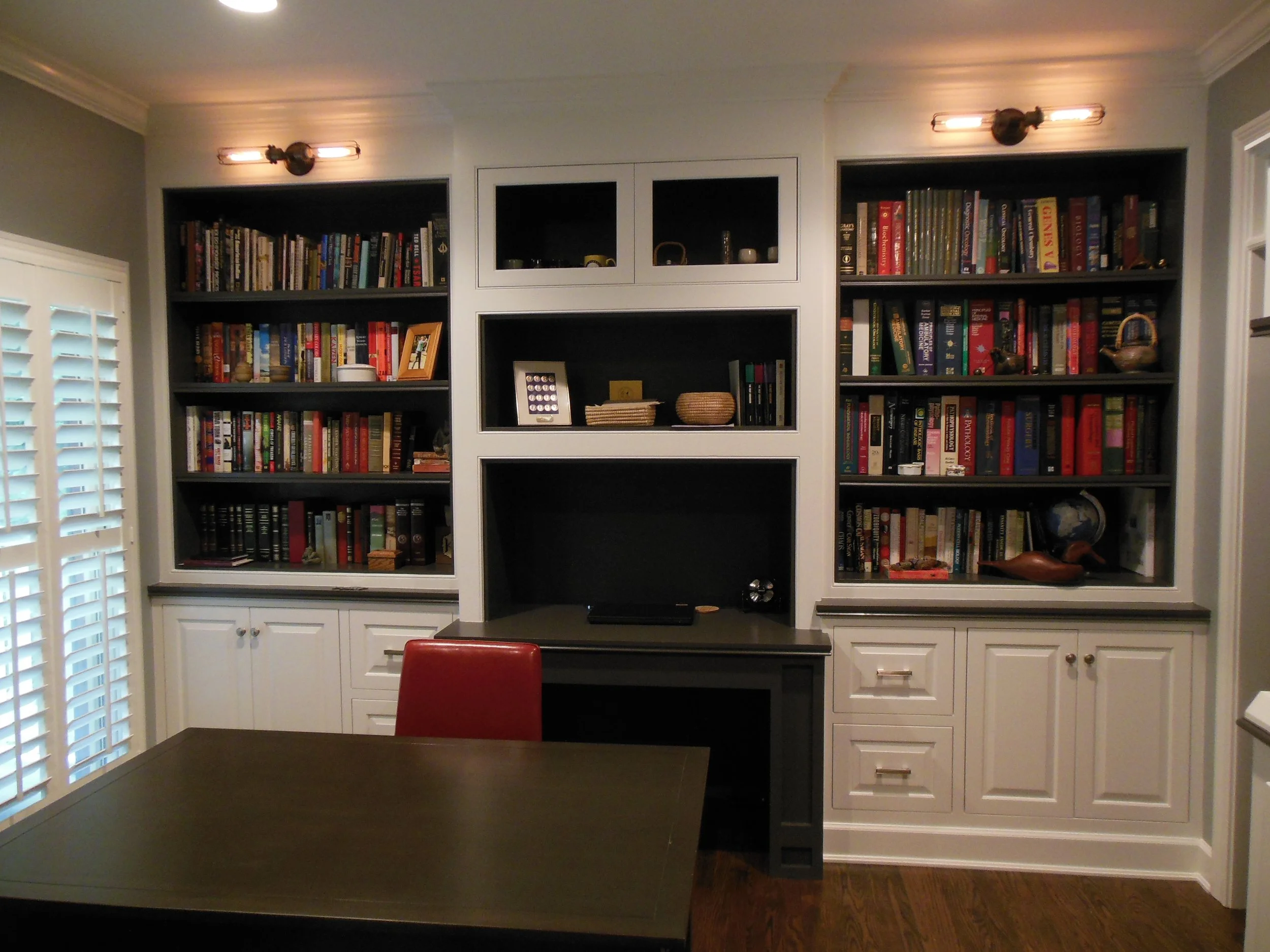 A room with built-in bookshelves filled with books on the wall behind a dark wooden table. White cabinet doors with silver handles are below the shelves, and a red chair is partially visible at the table. There are two modern wall sconces with expose