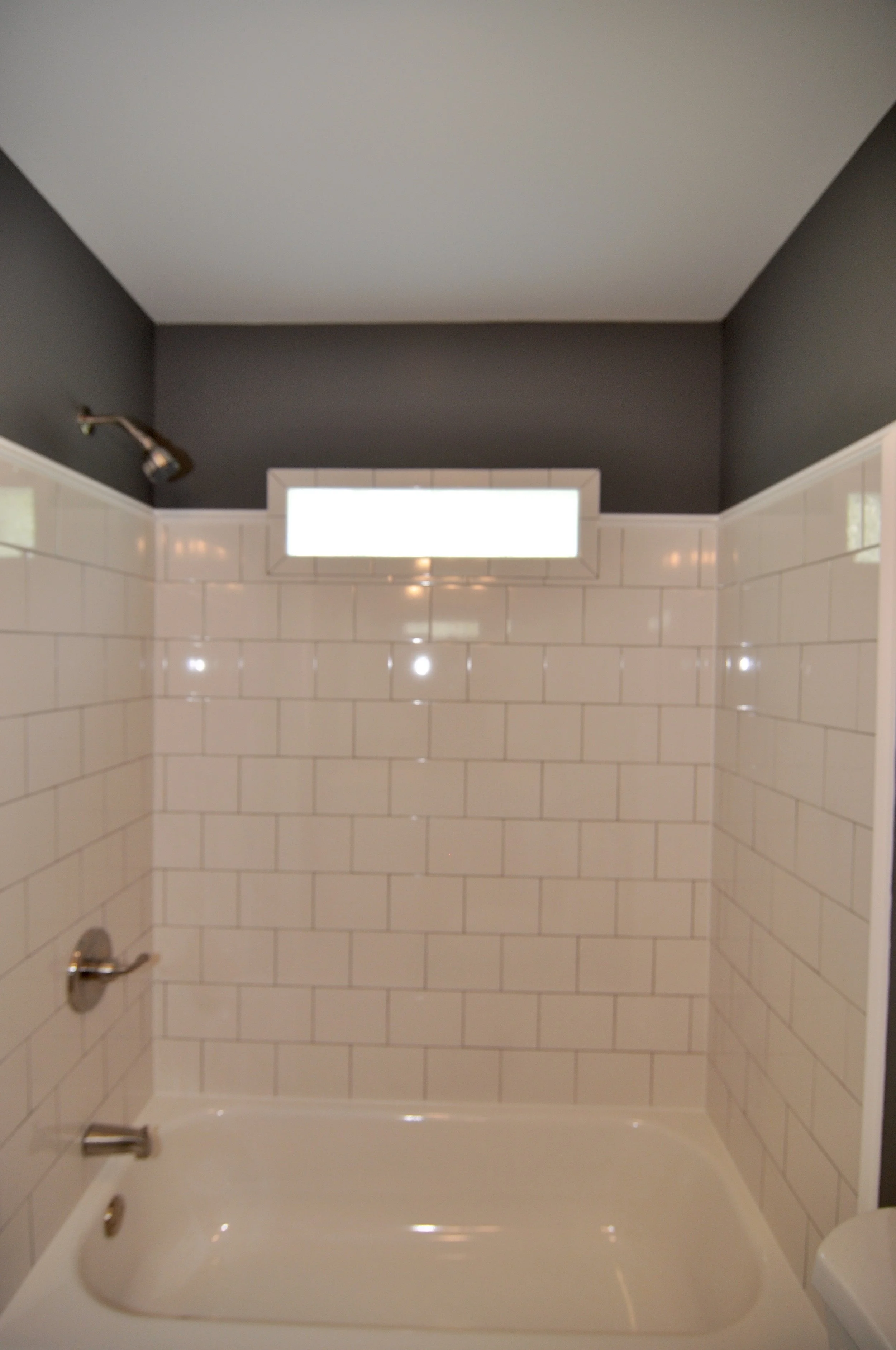 A bathroom with a white bathtub, cream tile surround, and dark gray painted walls. There is a showerhead in the upper left corner, a drain on the lower left, and a faucet on the lower right of the bathtub. A small rectangular window is on the wall ab