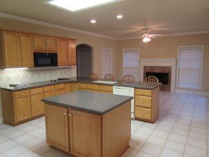 A before shot of a kitchen with wooden cabinets, a central island, and a dining area with chairs near a fireplace.