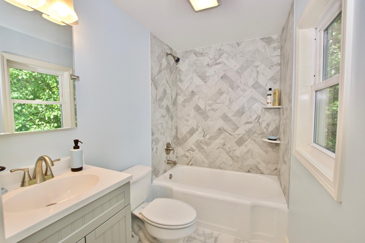 Bathroom with a vanity sink, mirror, toilet, bathtub with marble tile surround, and two windows showing greenery outside.