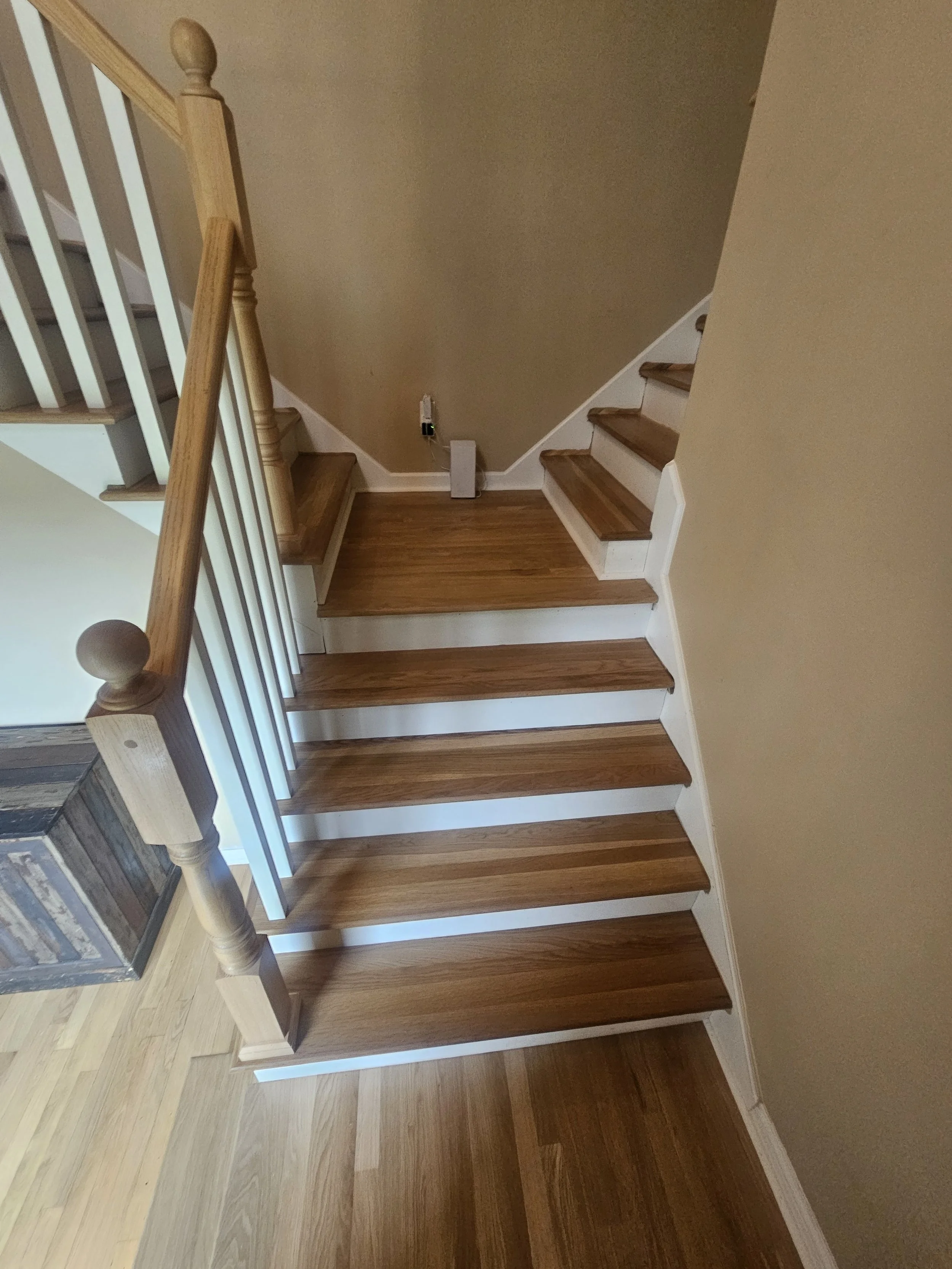 Photo of a staircase with wooden steps and a wooden handrail inside a house. The stairs are against a beige wall, and there is a small vent or electrical box at the bottom of the wall.
