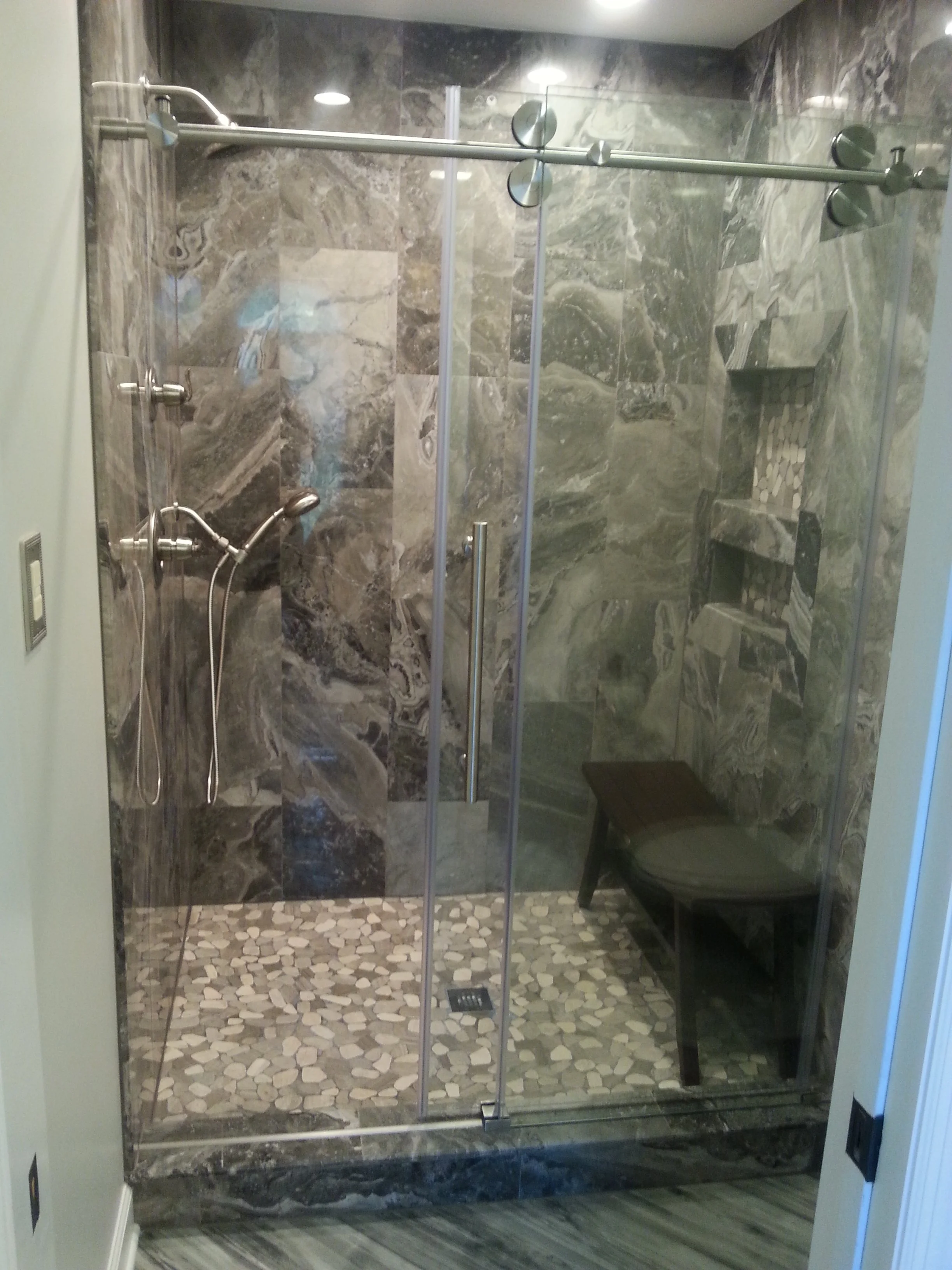 Modern walk-in shower with glass doors, marble walls, pebble tile floor, built-in niche, and a showerhead and handheld shower attached to the wall. There is a black bench inside the shower.