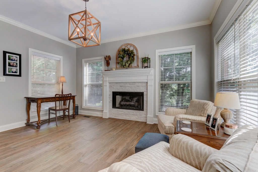 Living room with a fireplace, wooden floor, and large windows with blinds. There is a beige upholstered chair and a wooden side table with a lamp and framed photos. A small desk with a chair and a lamp is in the corner. A decorative wreath and wall art are above the fireplace, and a geometric light fixture hangs from the ceiling.