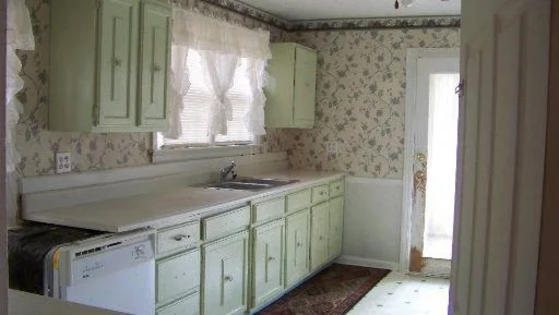 A before shot of a kitchen with light green cabinets, floral wallpaper, a window with lace curtains, a white countertop, and a washing machine at the end of the counter. There is a door leading outside or to another room.