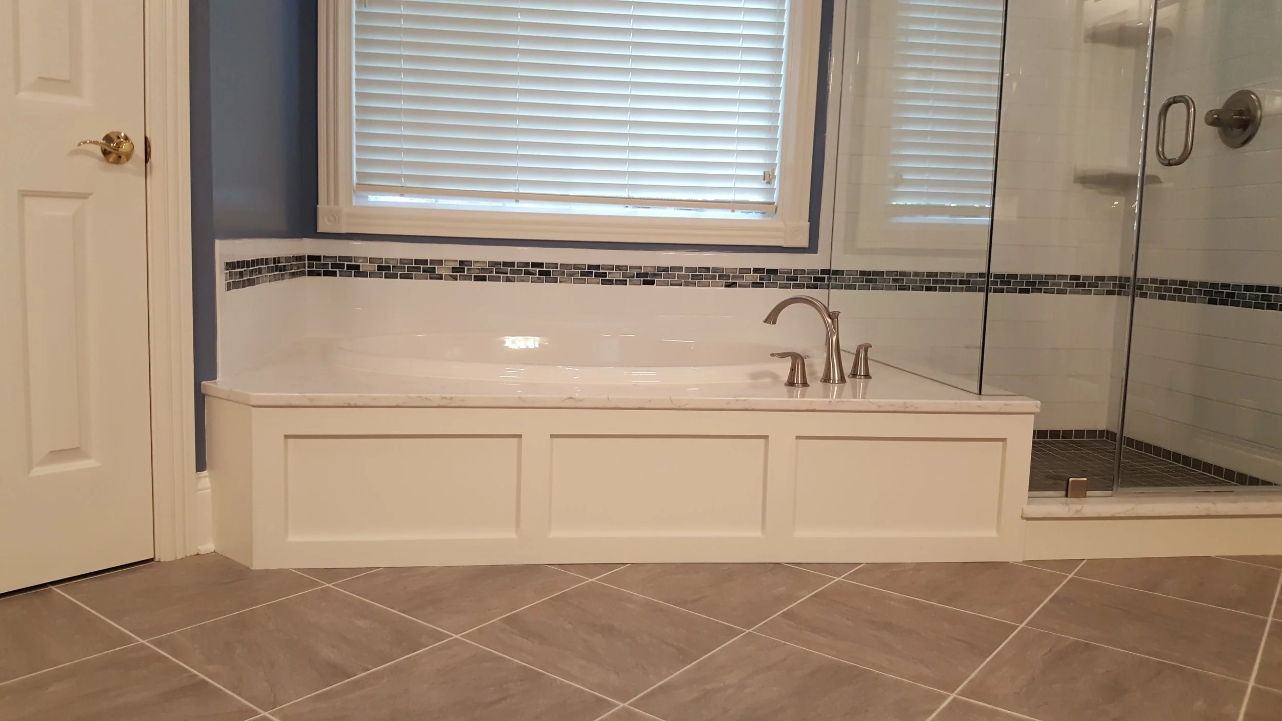 A bathroom with a built-in bathtub, a window with closed blinds, a glass-enclosed shower, and tan tile flooring.