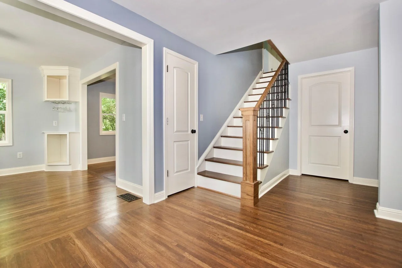 Interior of a house showing a staircase with wooden handrail and black metal balusters, hardwood floors, light blue walls, and white doors.