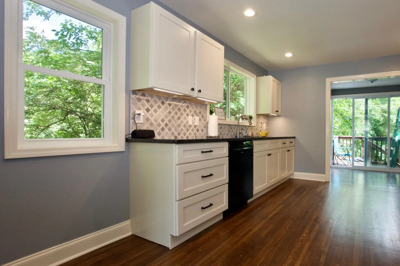 Kitchen with white cabinets, black countertops, gray patterned tile backsplash, wood flooring, and large windows showing green trees outside.