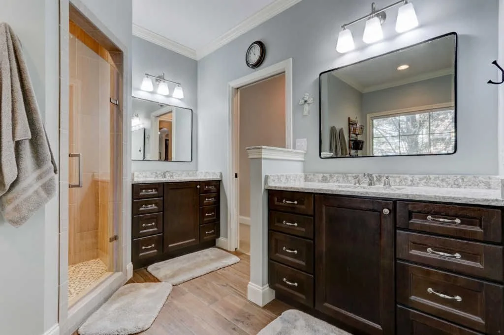 A bathroom with dark wooden cabinets and white marble countertops, large wall-mounted mirrors, a glass-enclosed shower with wood and tile interior, towels hanging on hooks, and a window showing trees outside.