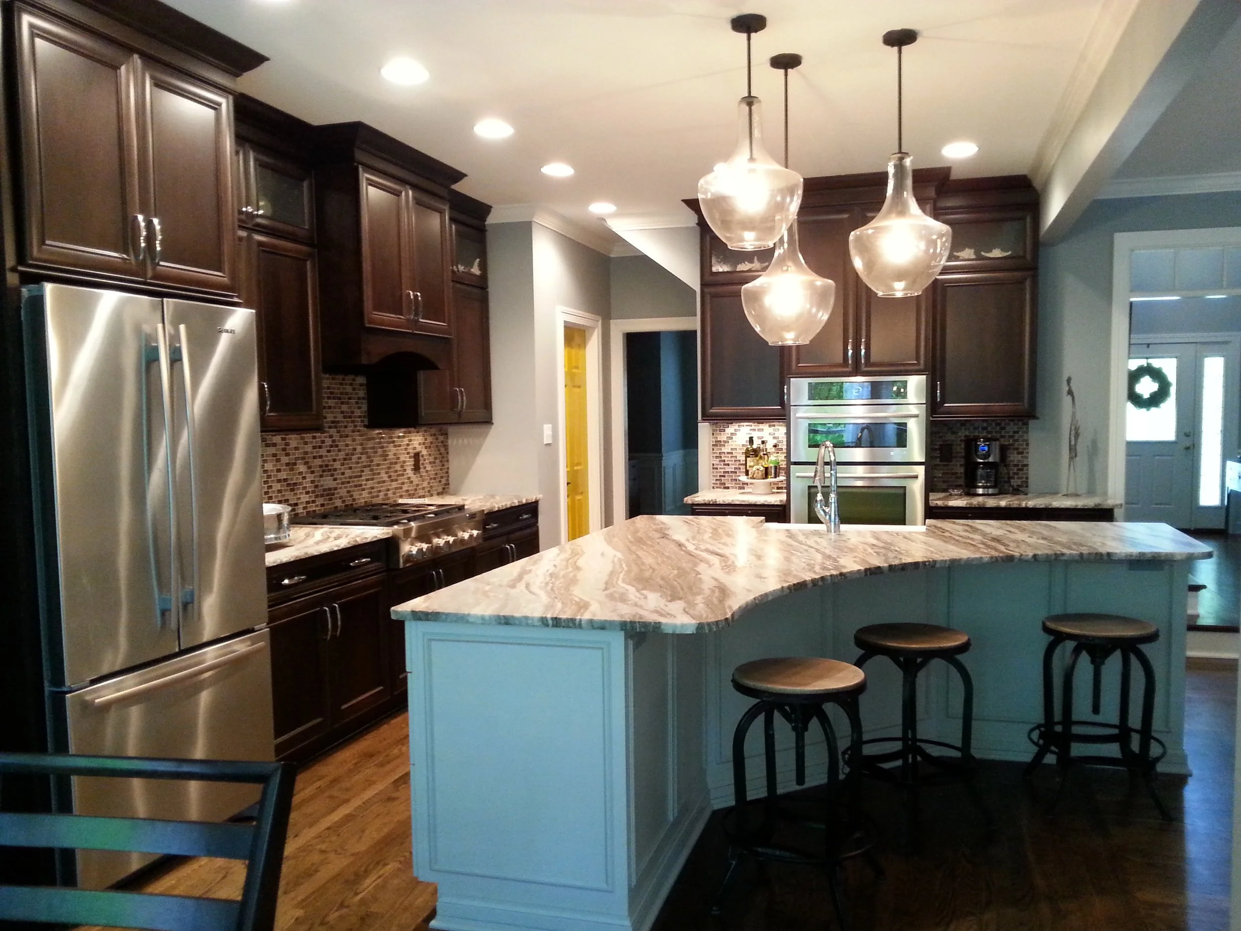 Modern kitchen with dark wood cabinets, stainless steel refrigerator, granite countertops, and a central island with a light blue base and bar stools with round wooden seats. Pendant lights hang above the island, and there is a mosaic tile backsplash
