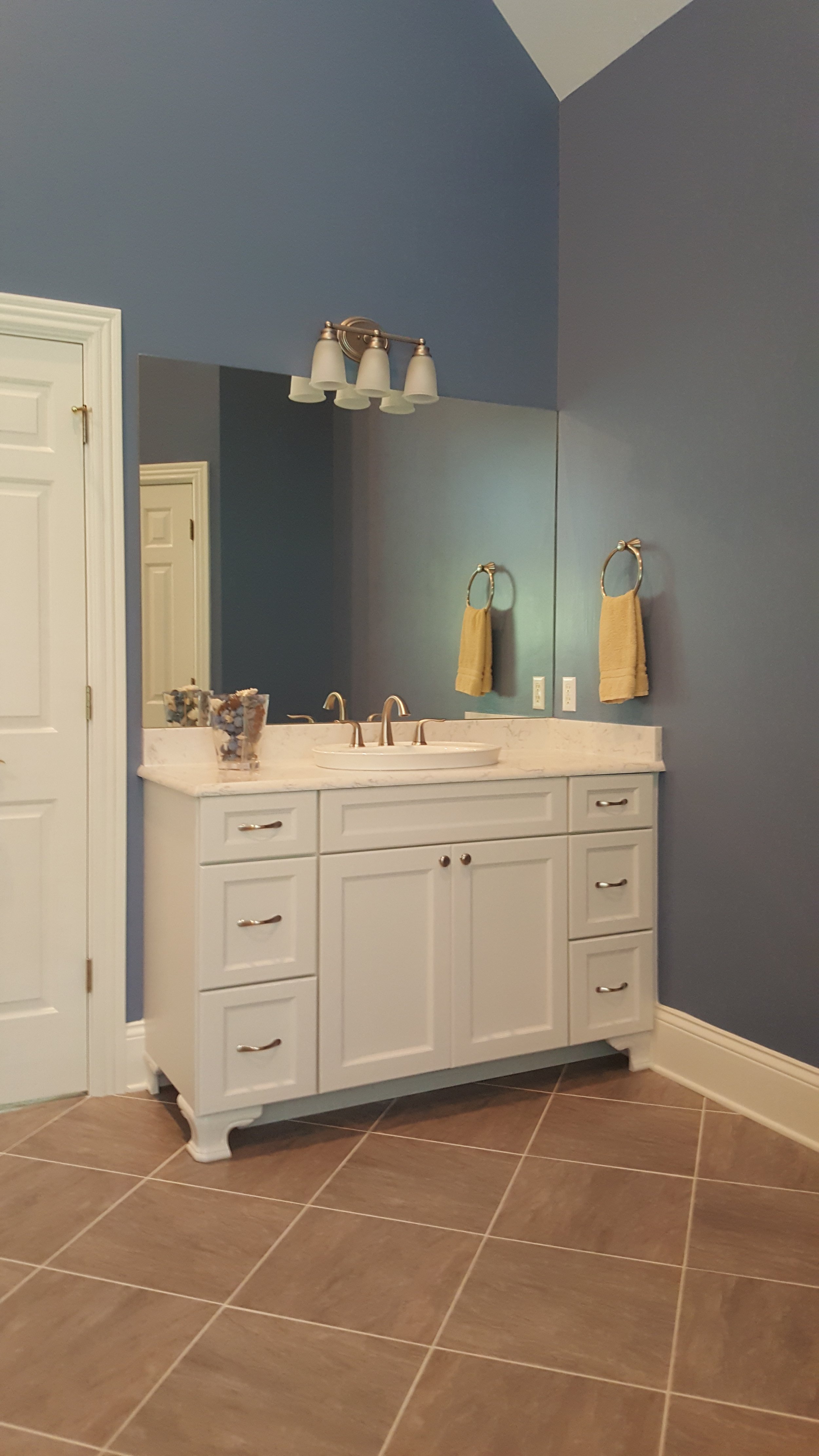 A bathroom vanity with a white cabinet, marble countertop, and a large mirror. There are two yellow towels hanging on the wall and a light fixture with four lamps above the mirror. The floor has large brown tiles.
