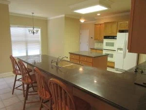 A before shot of an open kitchen with a long breakfast bar and three wooden chairs, white appliances, wooden cabinets, and a window with blinds