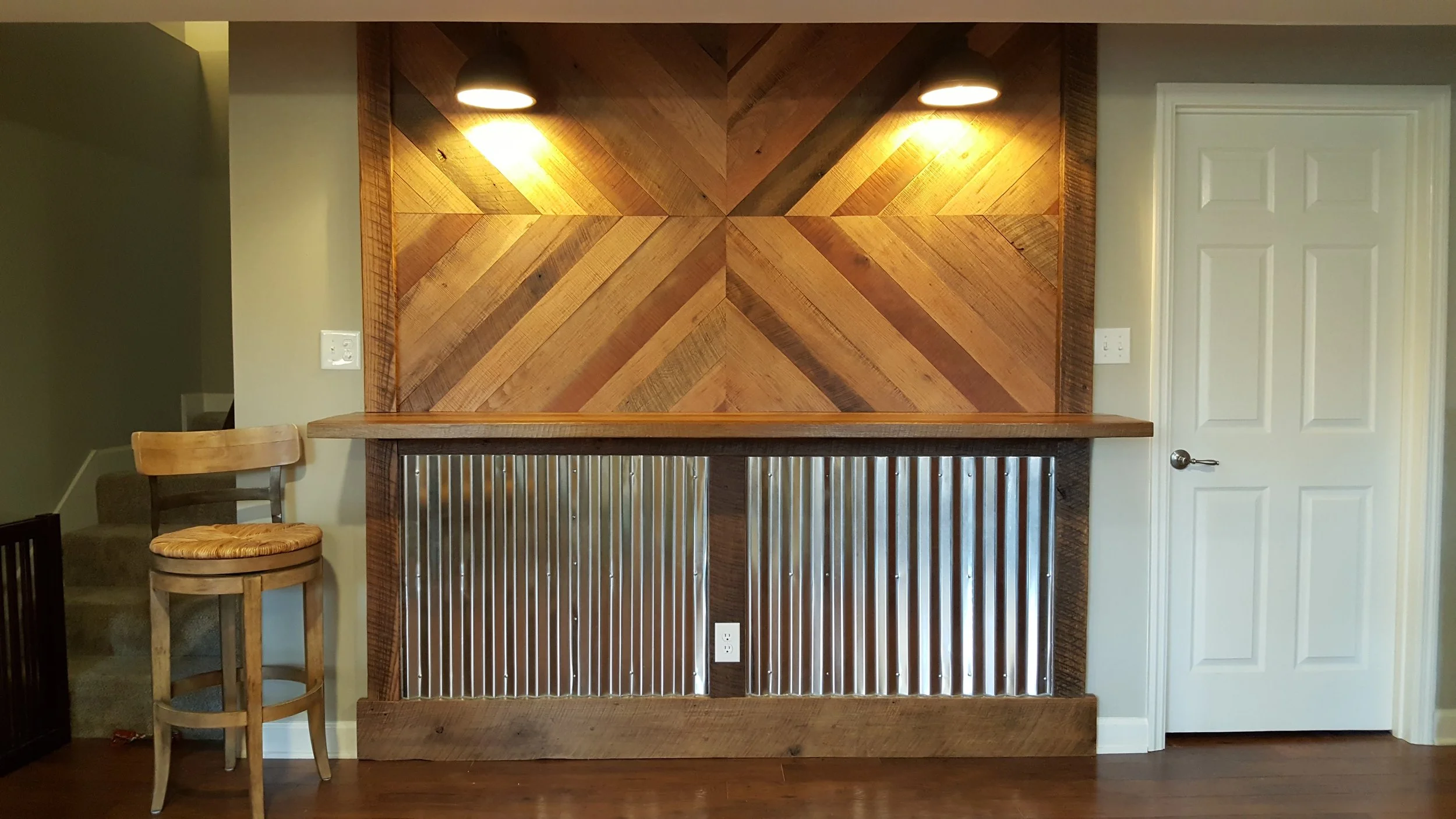 A wooden bar area with a chevron patterned wooden wall, two overhead lights, and a metal front panel. To the left, there is a wooden chair with a woven seat, and to the right, a white door.