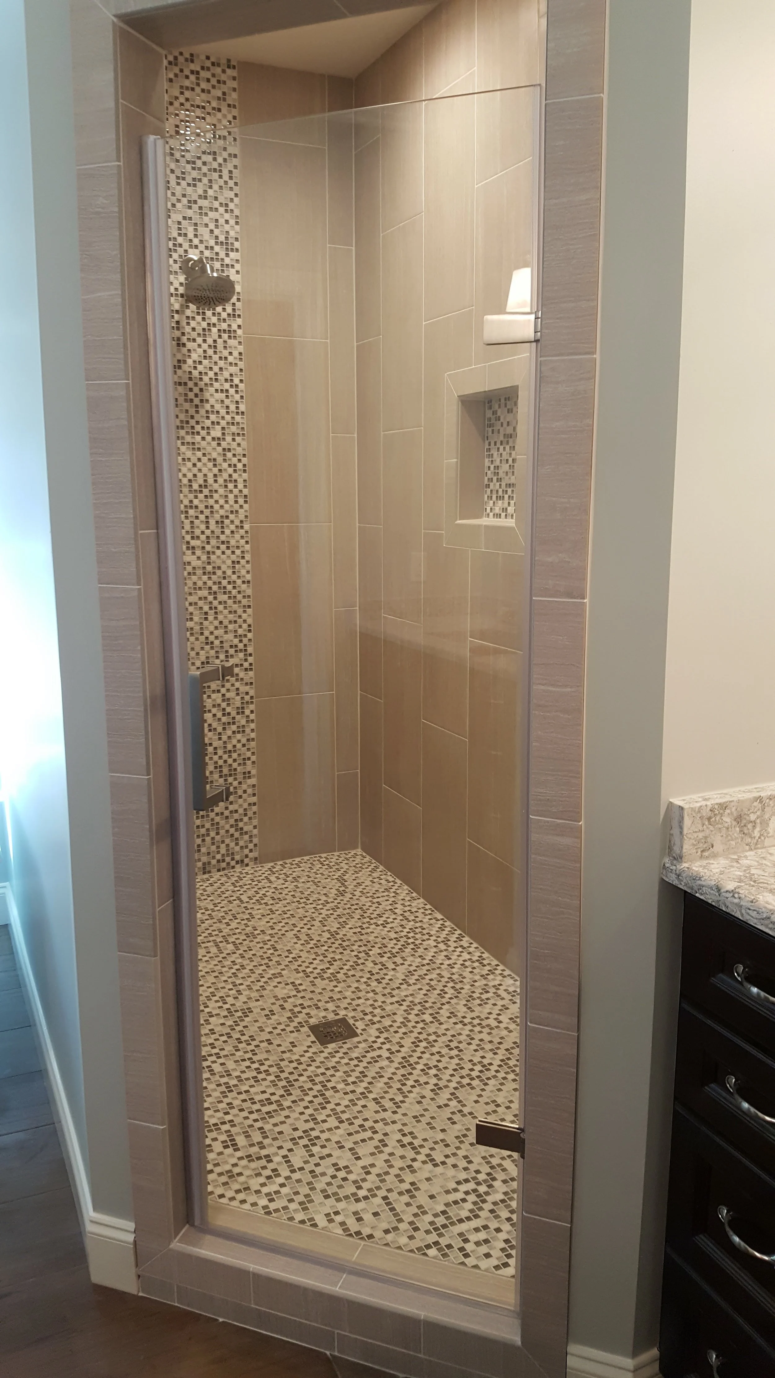 Bathroom shower with beige large tiles, a vertical strip of small mosaic tiles, a built-in niche with mosaic tiles, a rain shower head, and a glass door, next to a dark wooden cabinet with drawers.