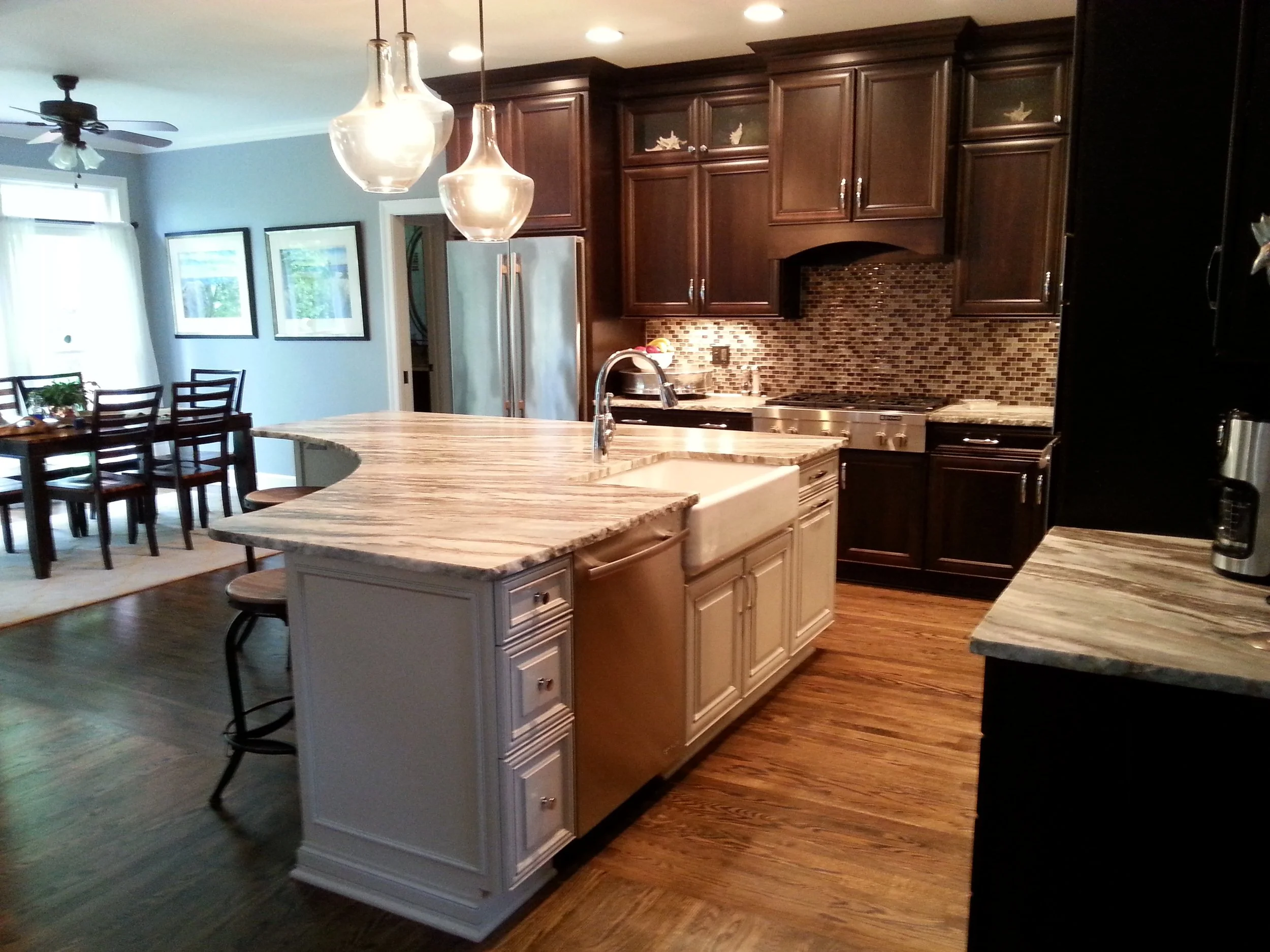 A kitchen with dark wooden cabinets, a mosaic tile backsplash, and a marble countertop island. There are pendant lights hanging above the island, a stainless steel refrigerator, and a stove. Adjacent is a dining area with a table and several chairs, 