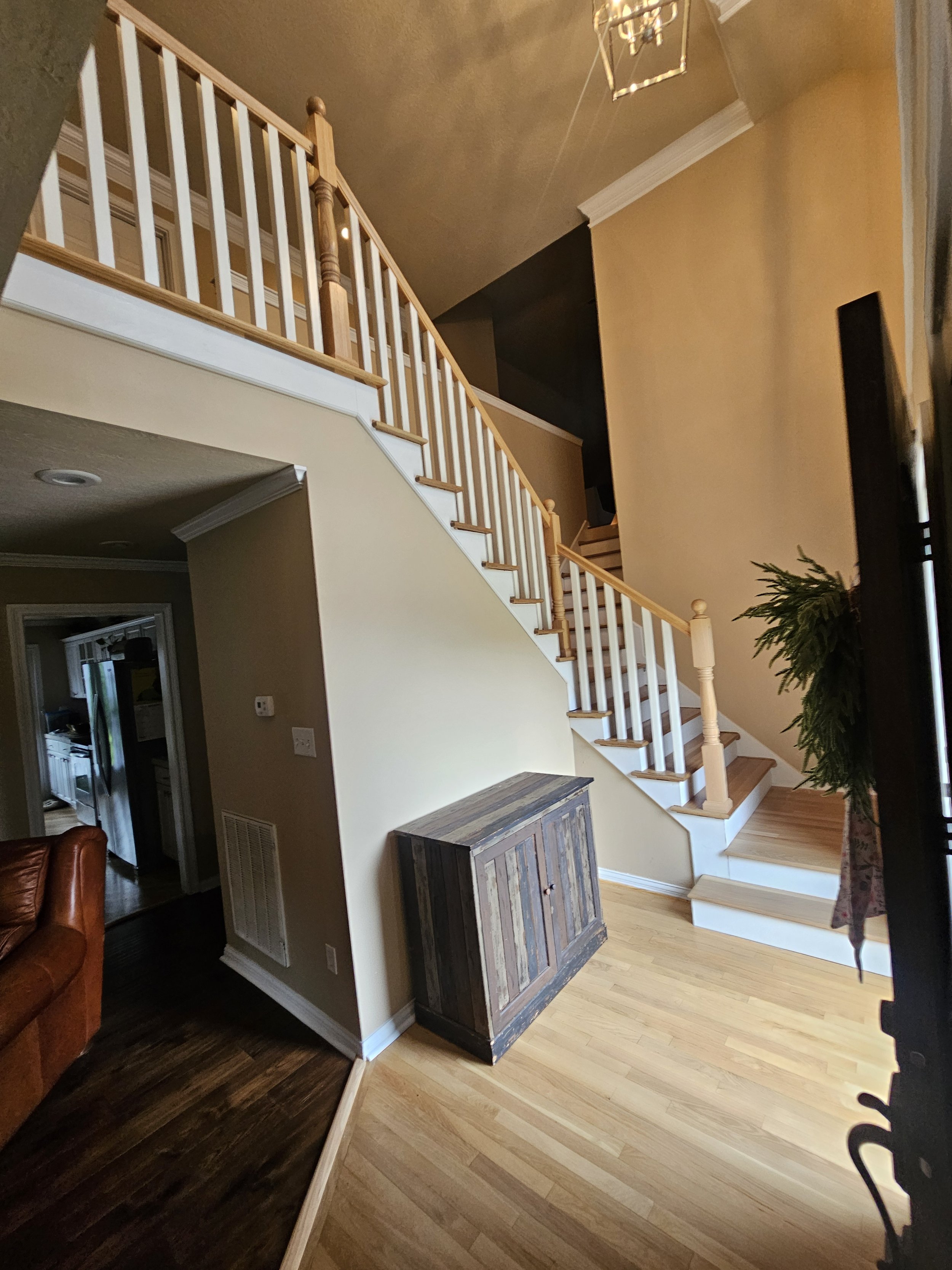 Interior of a house showing a staircase with a wooden railing, a dark and light wooden flooring, a beige wall, a black wall, a hanging ceiling light, a large potted plant, a wooden cabinet, and a doorway leading to another room.