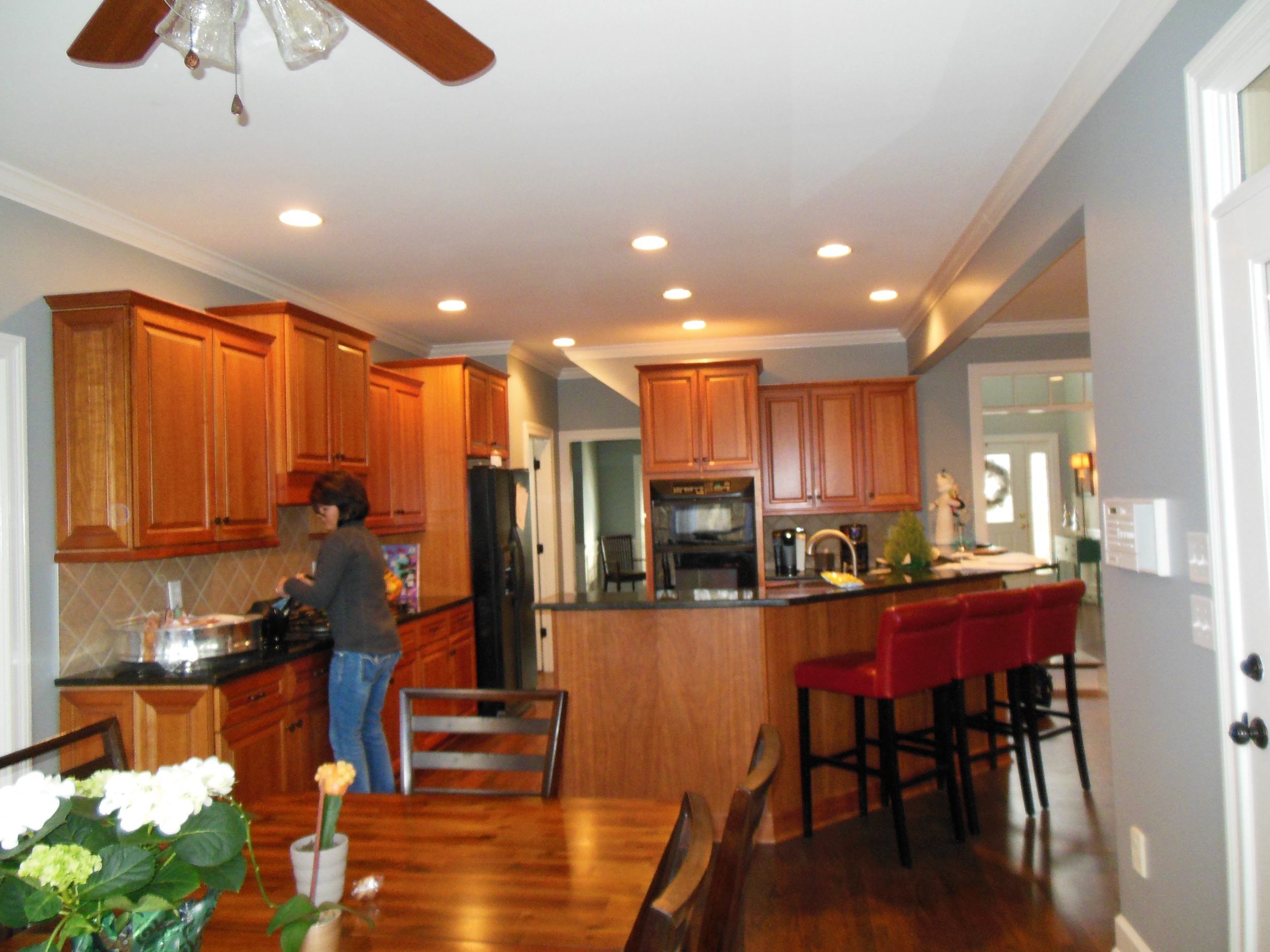 A before shot of a  kitchen with wooden cabinets, a round dining table with a flower arrangement, red chairs, and a ceiling fan with lights.