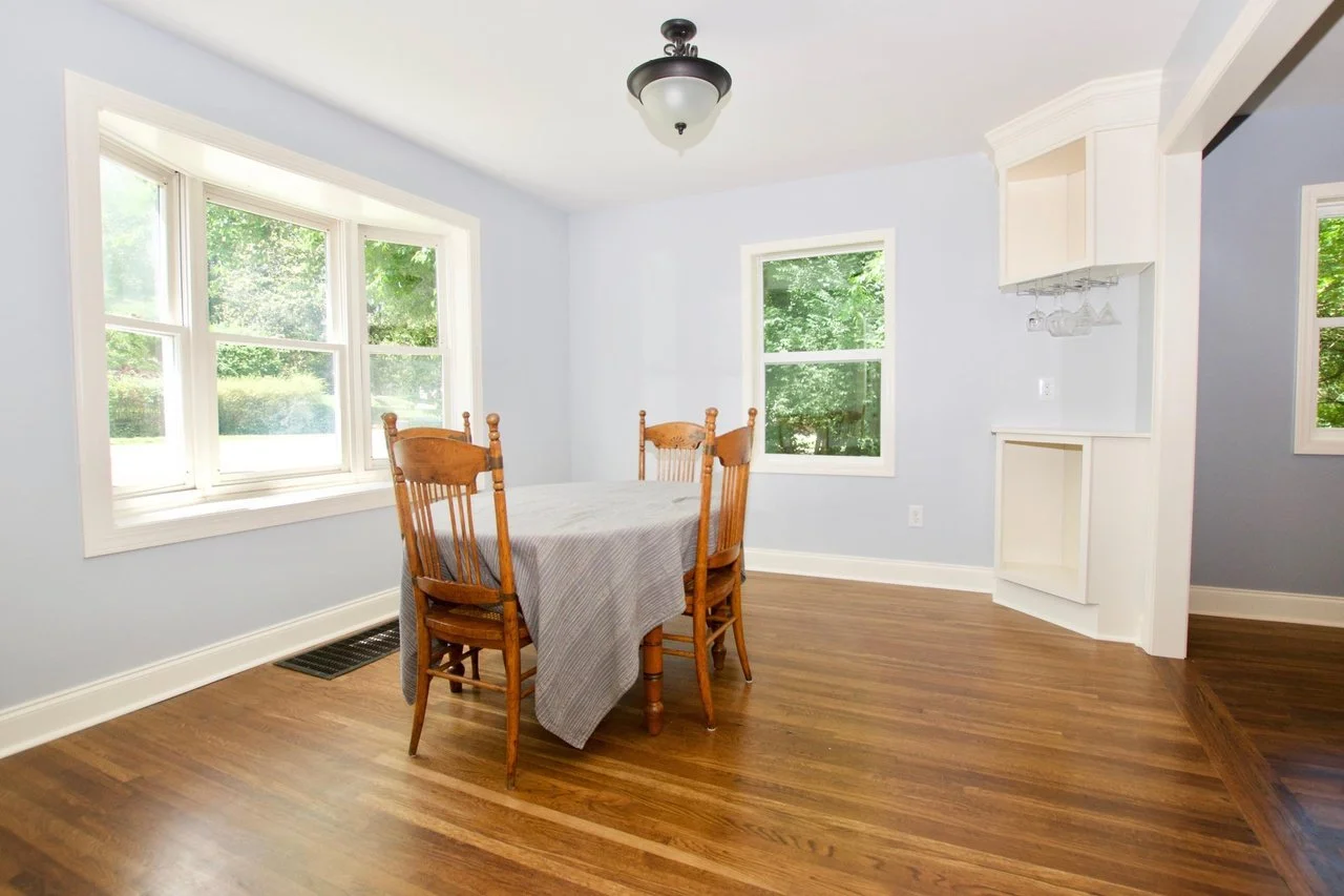 Dining room with wooden table and four chairs, light blue walls, hardwood floors, large windows with green outdoor view, ceiling light fixture, built-in cabinet with hanging glasses.