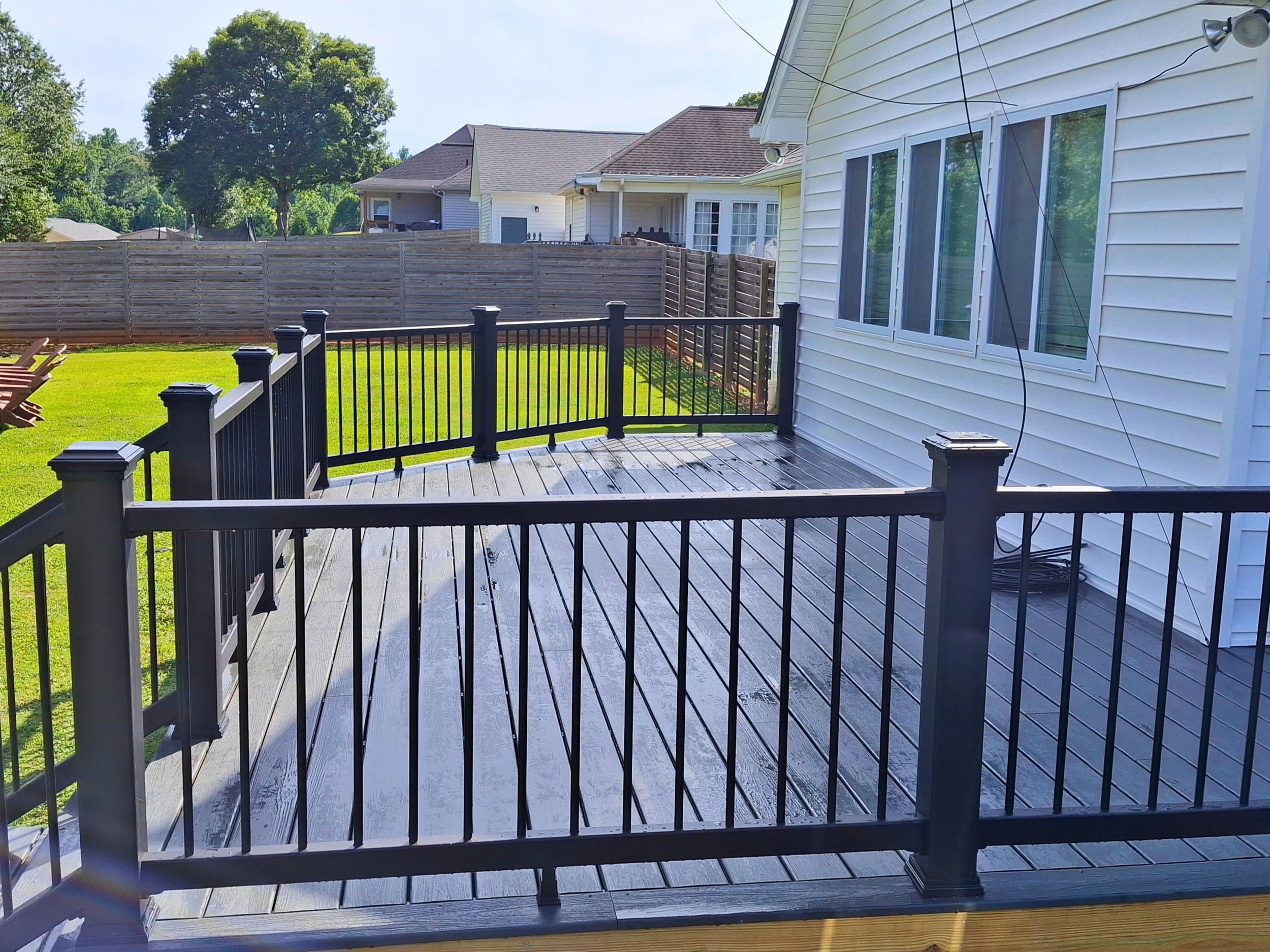 View of a backyard with a deck, black railing, lawn, wooden fence, neighboring houses, large trees, and clear sky.