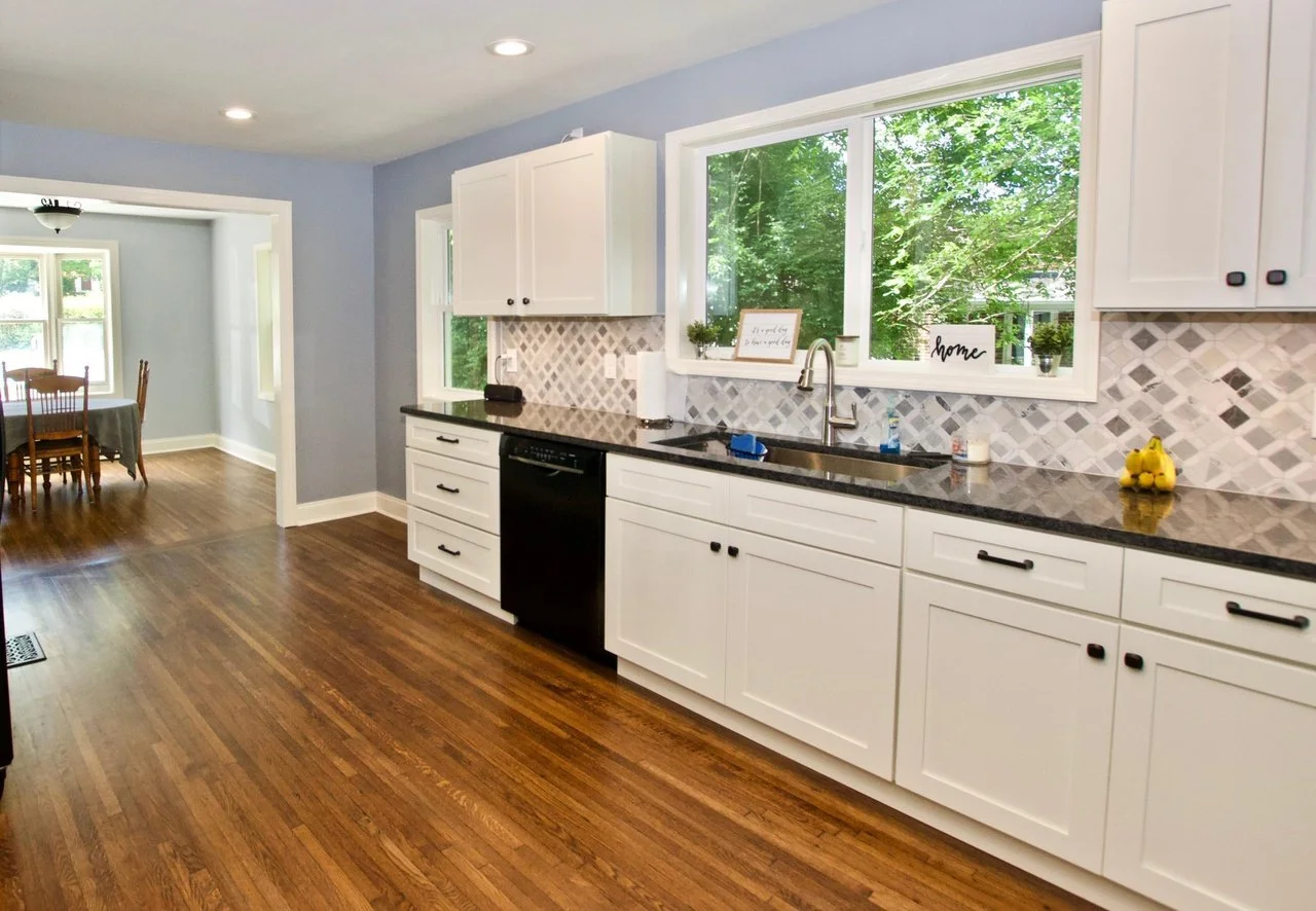 Kitchen with white cabinets, black countertops, diamond-patterned backsplash, large window showing green trees outside, hardwood floor, and a view into a dining area with a table and chairs.