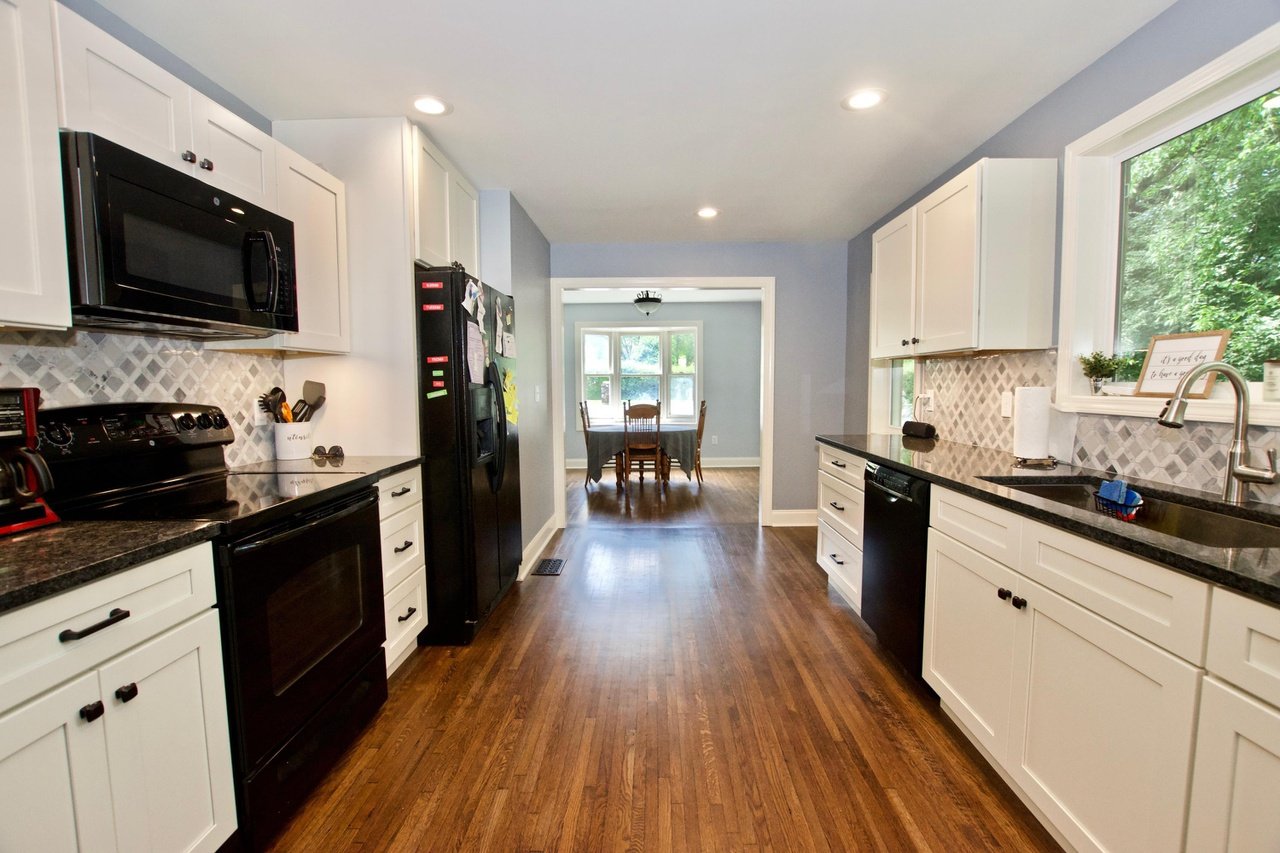 A kitchen with white cabinets, black appliances, and hardwood floors, leading to a dining area with a round table and chairs near windows.